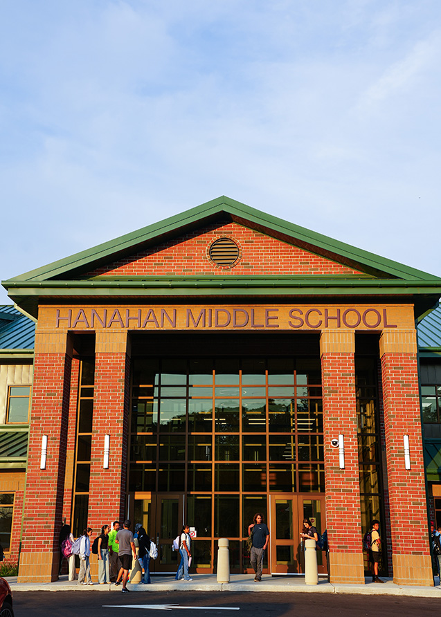 A school building with a triangular roof, glass doors, and brick walls. The sign reads "HANAHAN MIDDLE SCHOOL." Several people are standing outside.