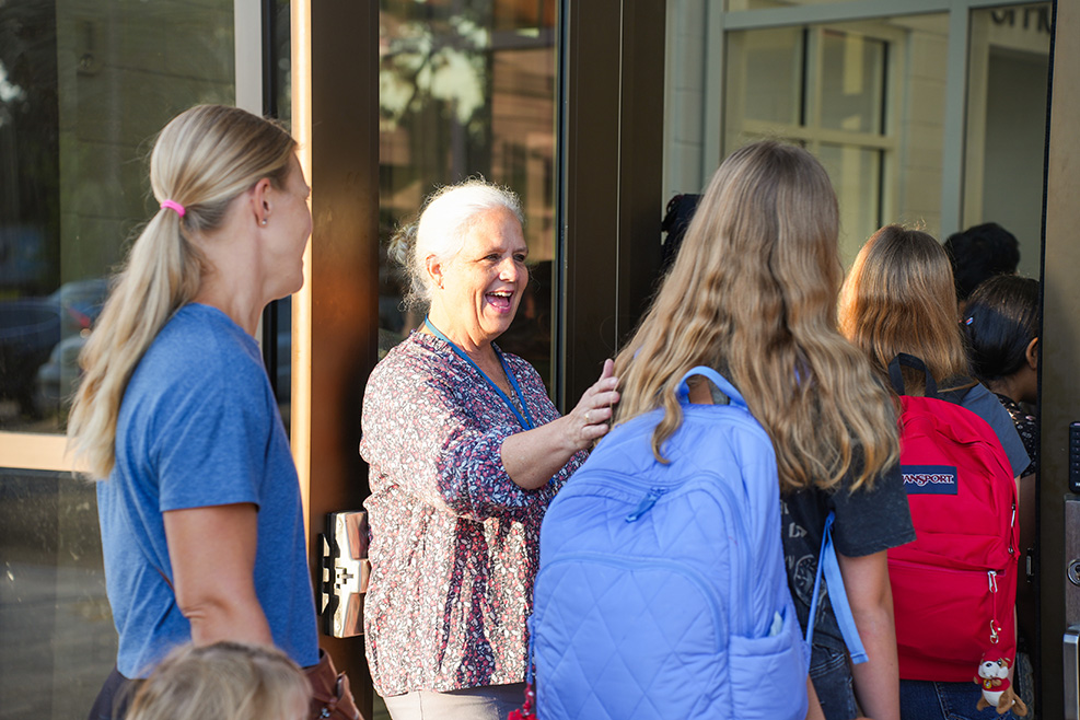 Women and girls with backpacks stand outside a glass door, with sunlight visible through it.