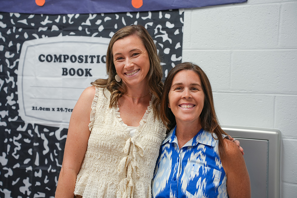 Two women smile next to a wall, one wearing a white top, the other a blue and white shirt.