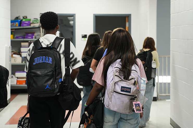 Students walking in a hallway. Some have backpacks. A shelf with various items in the background.