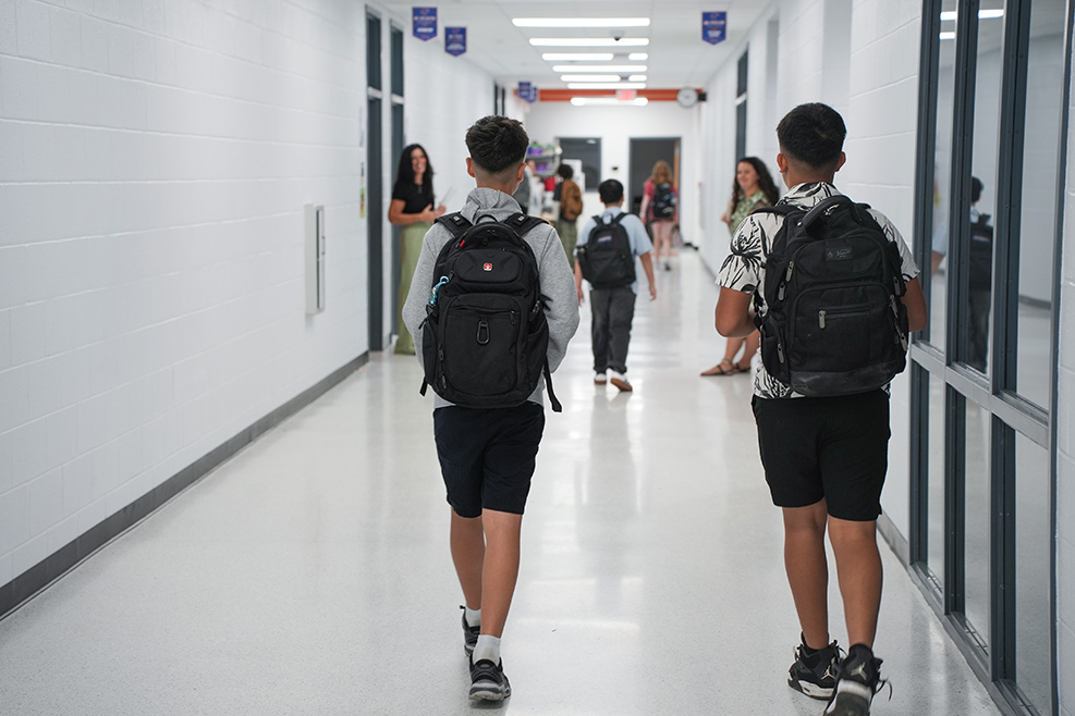 A group of students wearing backpacks walk in a school hallway with white walls and glass windows.
