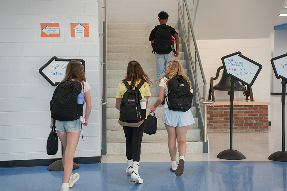 Four students in backpacks walk through a school hallway; one climbs stairs. White walls, blue floor, and signs.