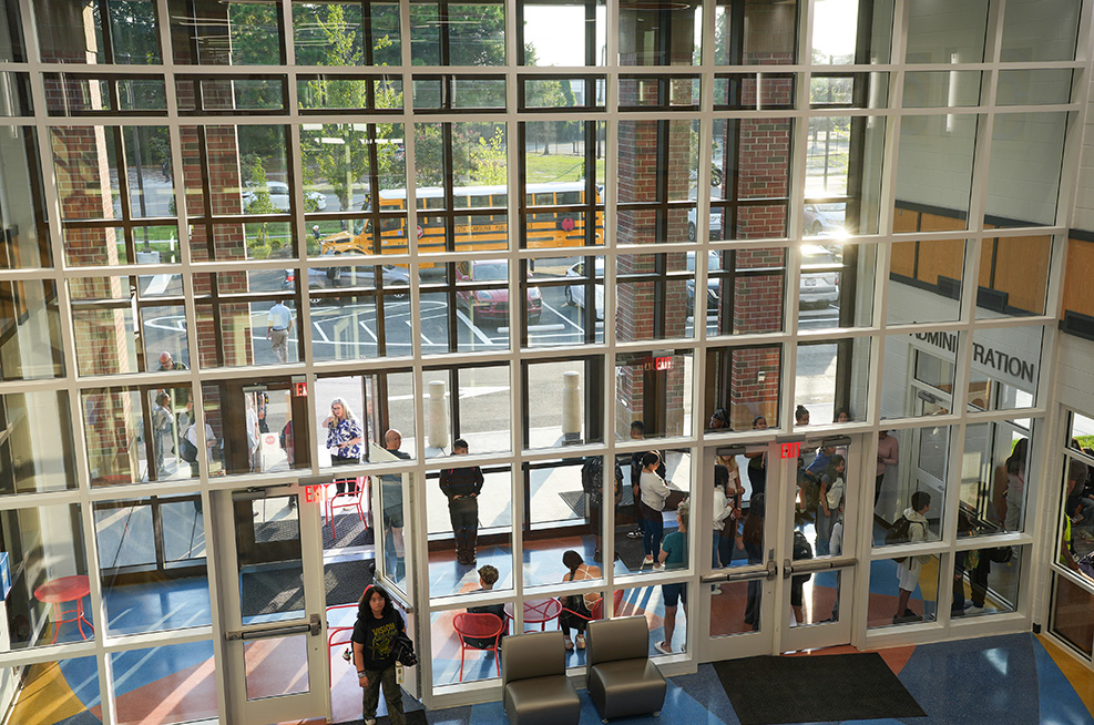A room with large glass windows shows people inside and a school bus outside on a sunny day.