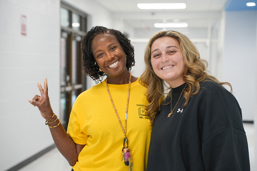 Two women stand in a brightly lit hallway, one in a yellow shirt and the other in black, both smiling.