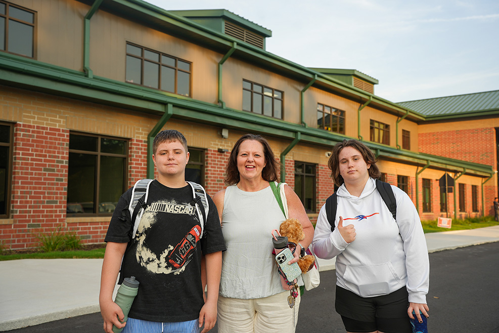 Three people stand in front of a brick building with green trim, smiling. Two are holding backpacks and one holds a stuffed toy.