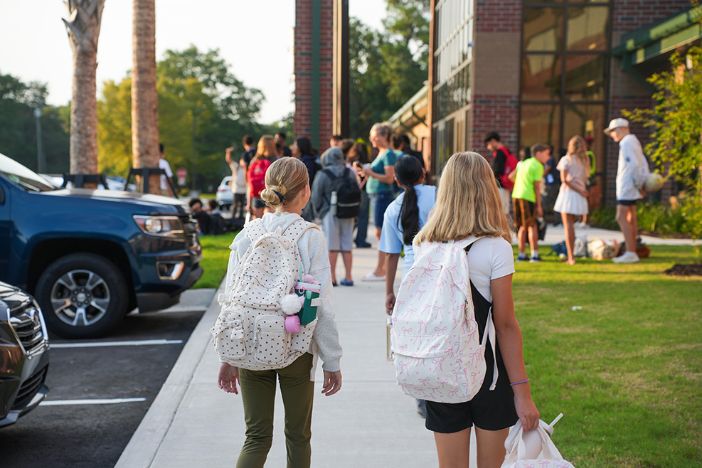 A group of students, some carrying backpacks, walks on a sidewalk near parked vehicles and buildings.