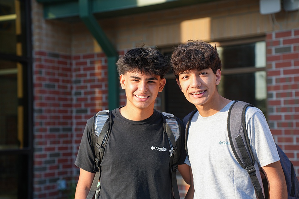 Two boys smiling, one in a black t-shirt and one in a white t-shirt, both with backpacks. They stand in front of a brick building.