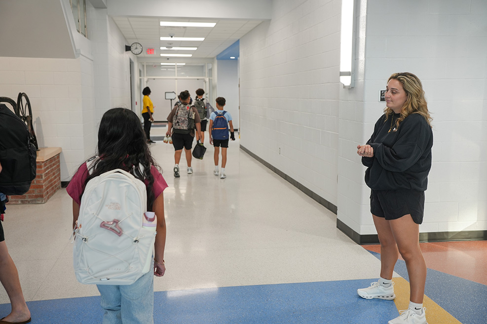 Hallway with white walls and blue flooring. A person in black converses with a girl in white and blue.