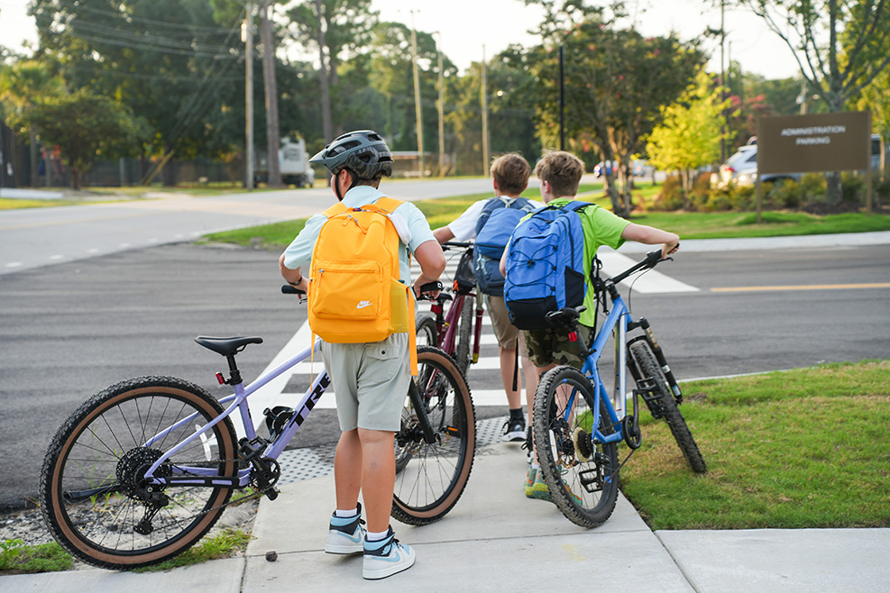 Three children in casual clothing wait at a sidewalk with their bikes. One wears a helmet.