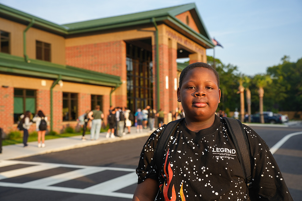 A child stands in front of a school building with brick walls and green roofing. People walk behind.