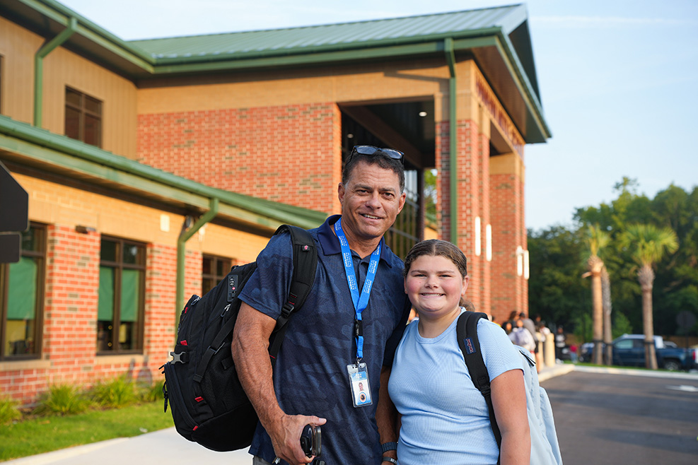A man and a young girl stand outside a brick building. The man has a backpack on.