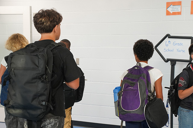 Students stand near a hallway, some carrying backpacks. One holds a sign with directions.