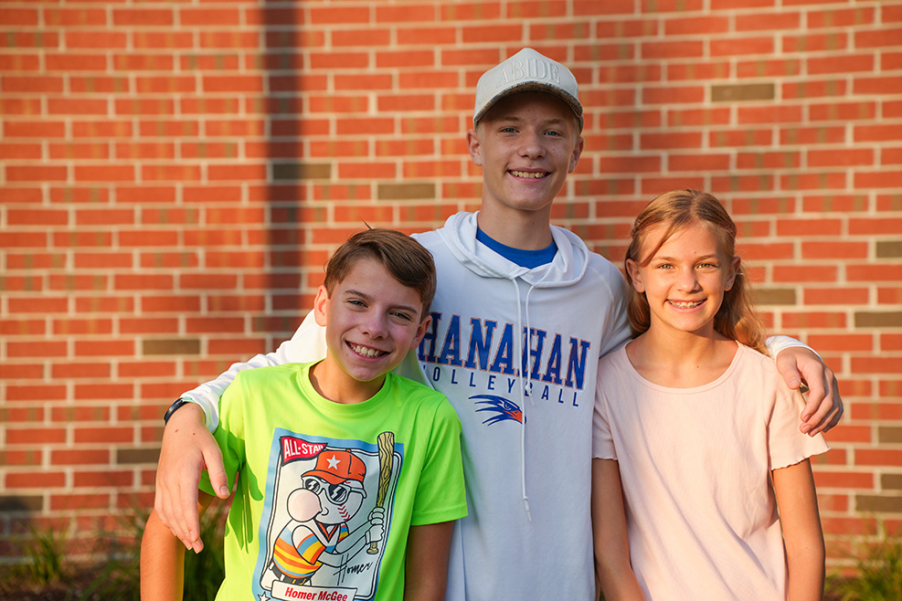 Three children in front of a red brick wall, one in a green t-shirt, one in a white hoodie, and one in a pink shirt.