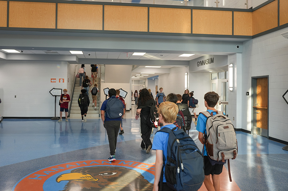 A hallway with white walls and blue flooring. Students are walking, carrying backpacks. A circular emblem with an eagle sits on the floor.