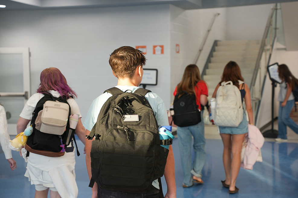 Five individuals walk in a hallway, wearing backpacks; one has colorful hair. Behind them, stairs lead upwards.
