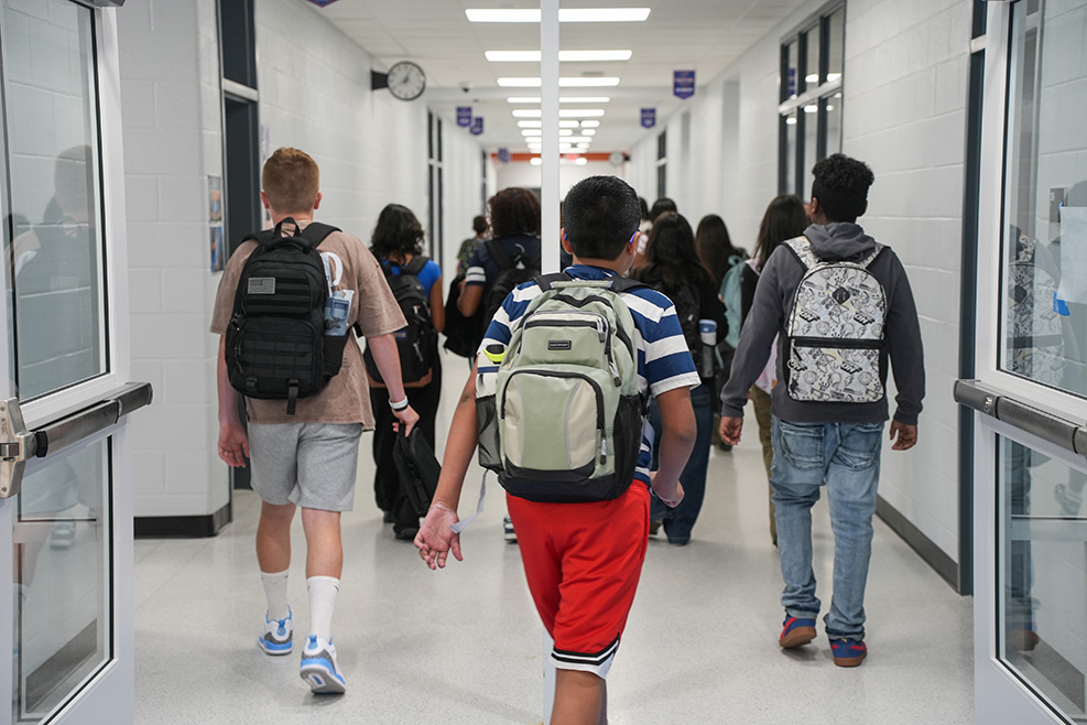 Students walking down a school hallway with backpacks. Bright fluorescent lighting. Two people walking hand-in-hand.