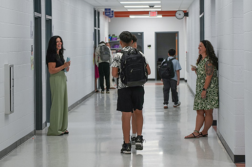 People stand and walk in a white hallway with doors. A woman in a green dress holds a drink.