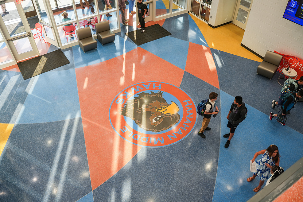 A top-down view of a hallway with a logo on the floor. Several people are walking around.