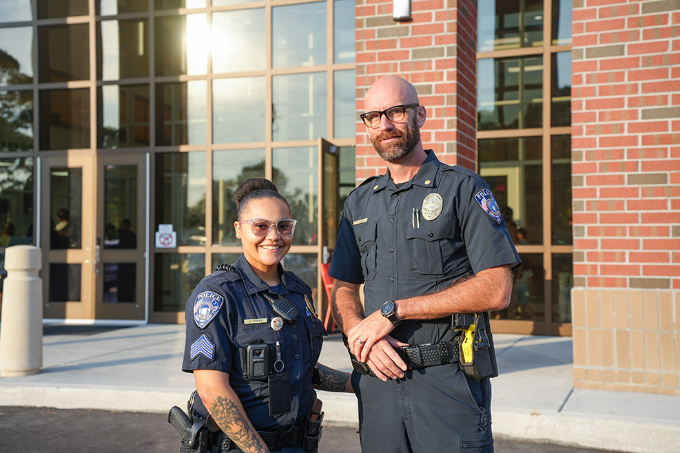 Two police officers stand before a brick building with large windows. The officer on the right wears glasses.