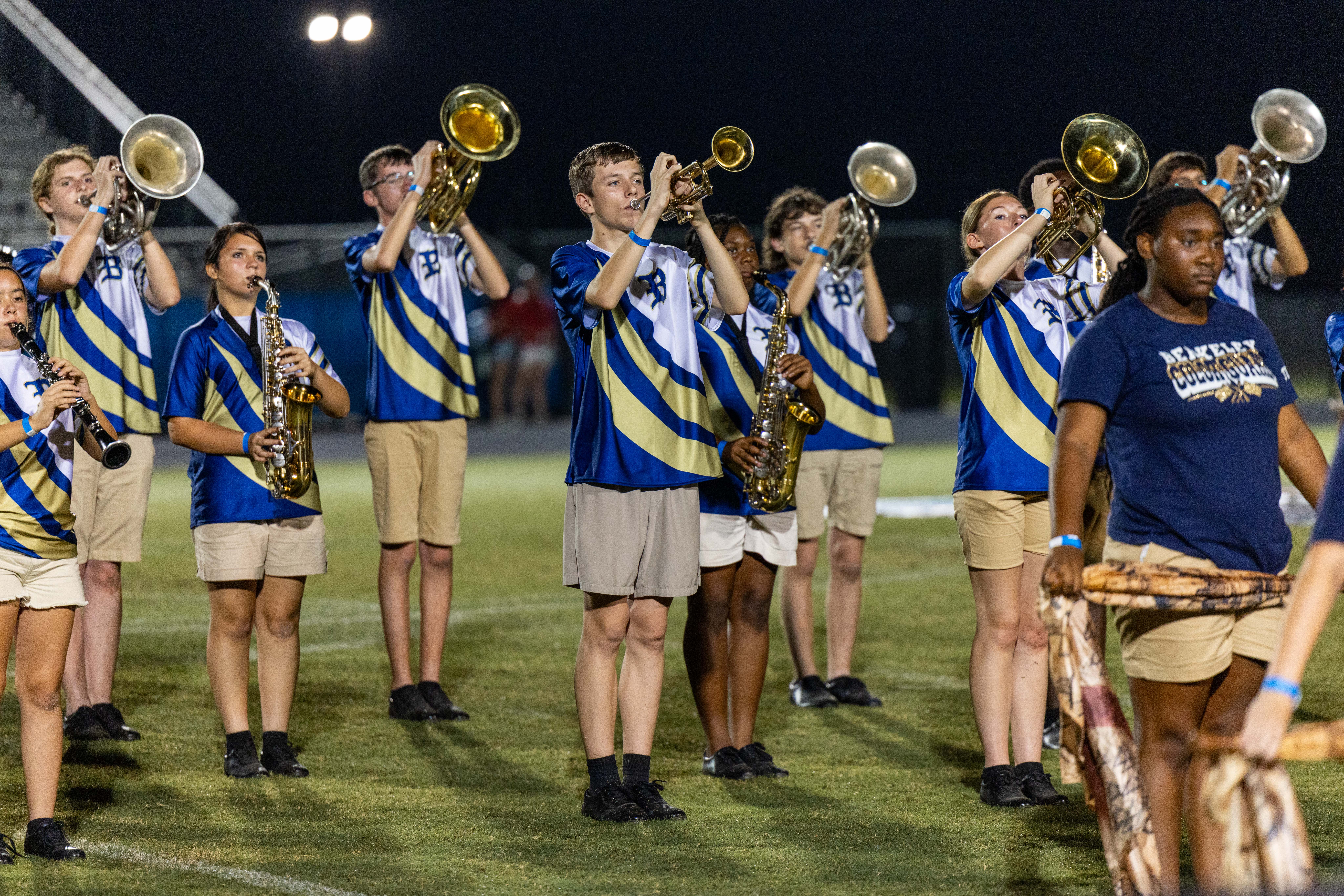 A group of musicians dressed in blue and white uniforms play instruments on a field at night.