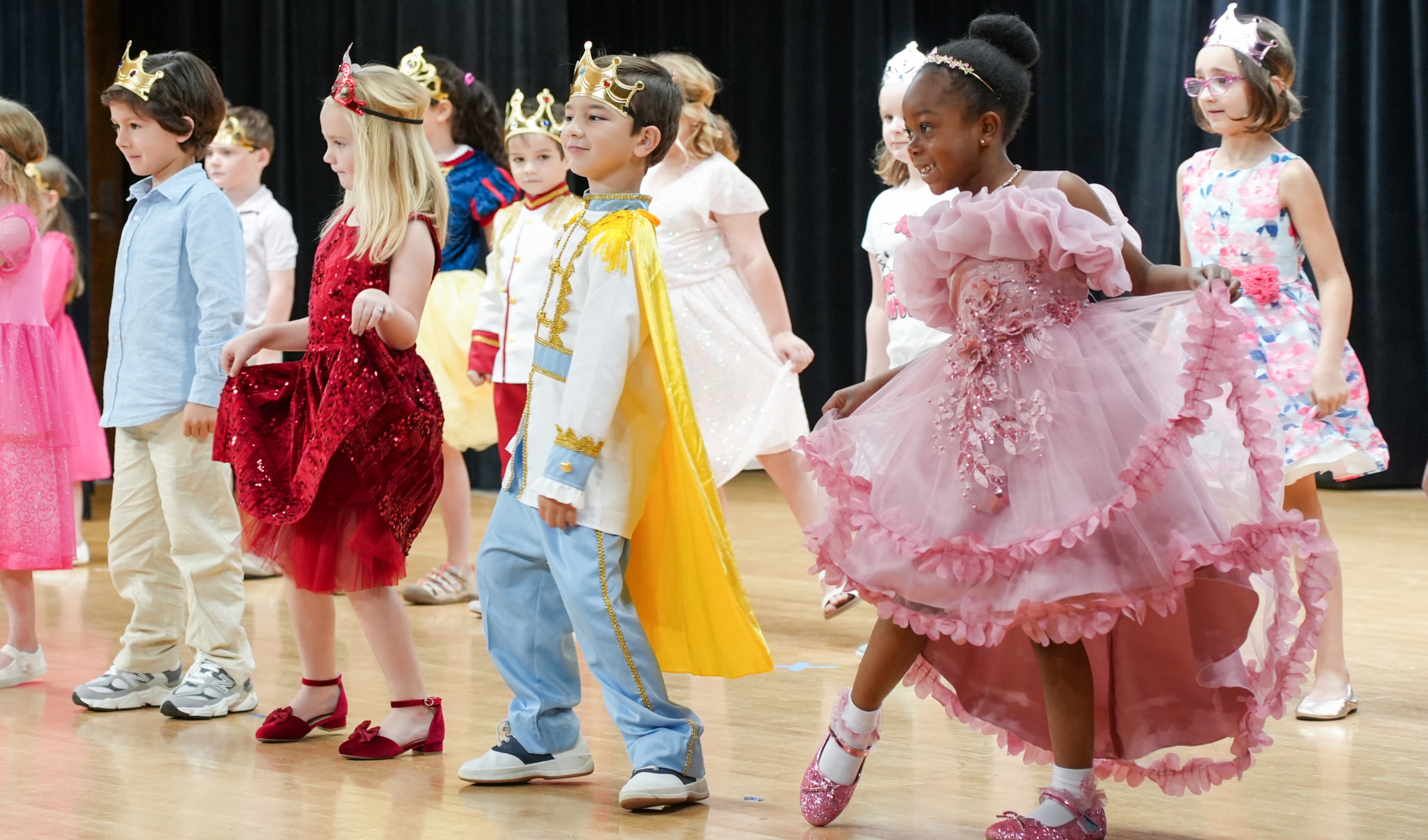 A group of children in various costumes dance on a wooden floor. Some wear crowns, capes, and shoes.