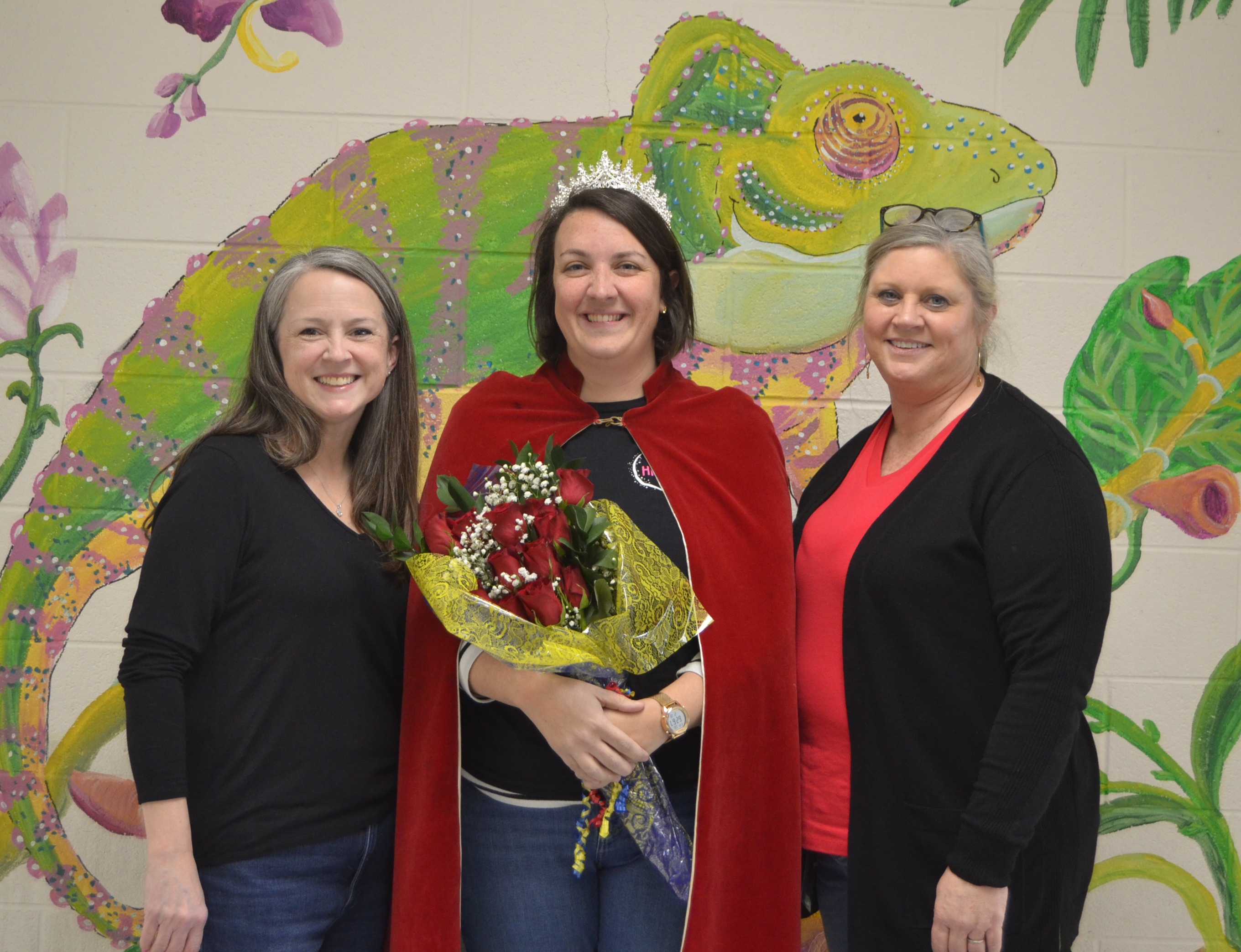Three women stand together in front of a colorful wall mural with a chameleon and tropical plants.