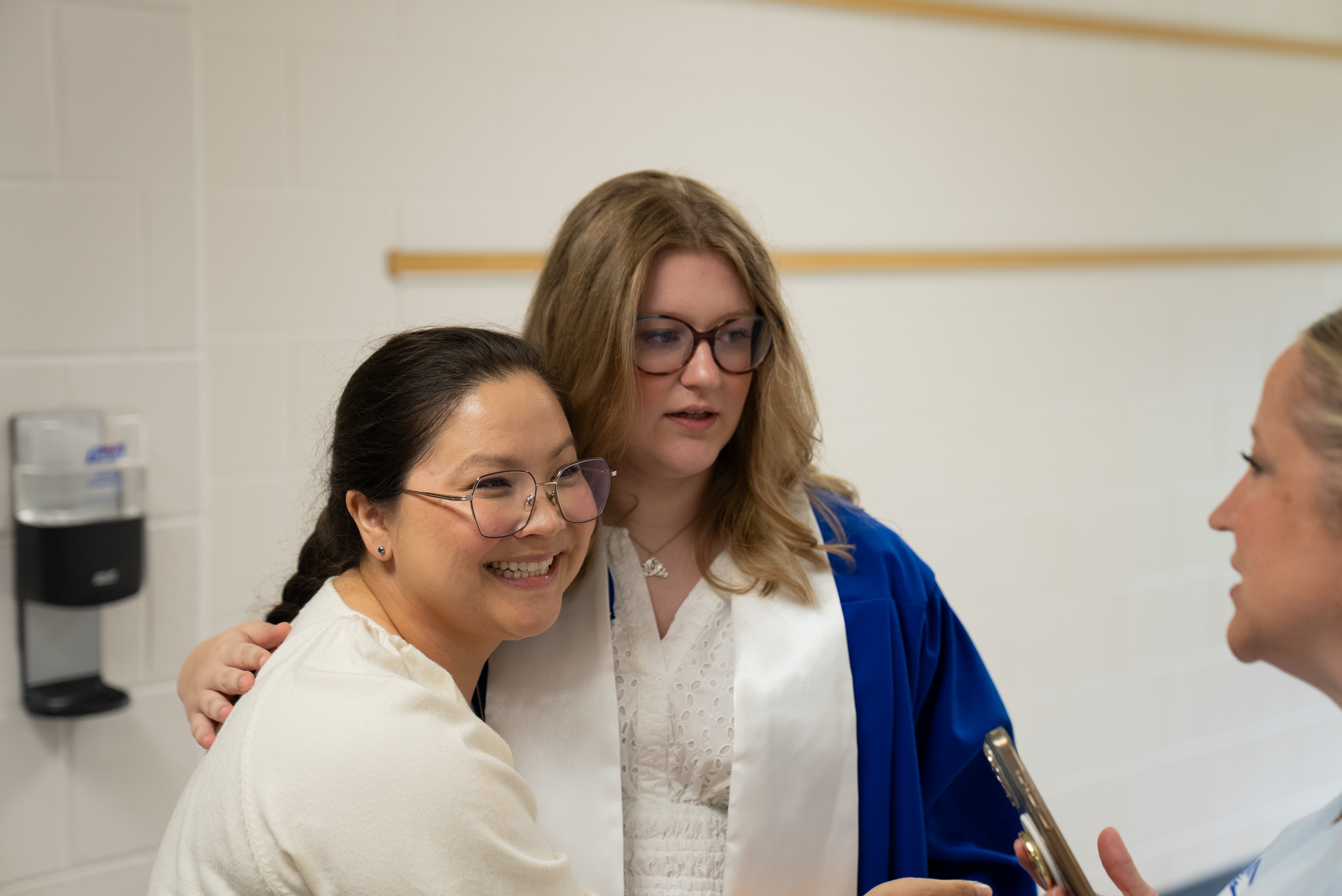 Three women stand in a room; one with a blue robe and necklace, another in a white shirt, and a third with a trombone.