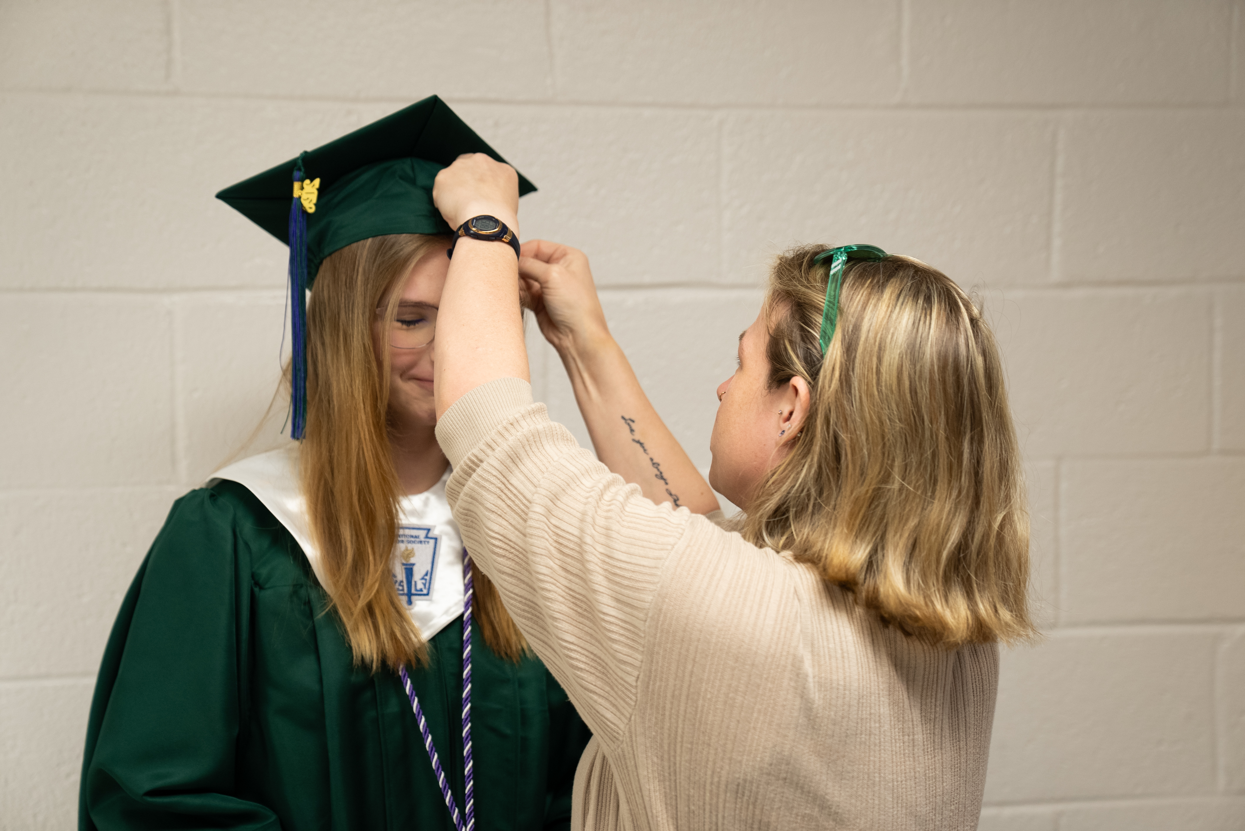 A woman in a graduation gown adjusts the cap of another woman in front of a white wall.