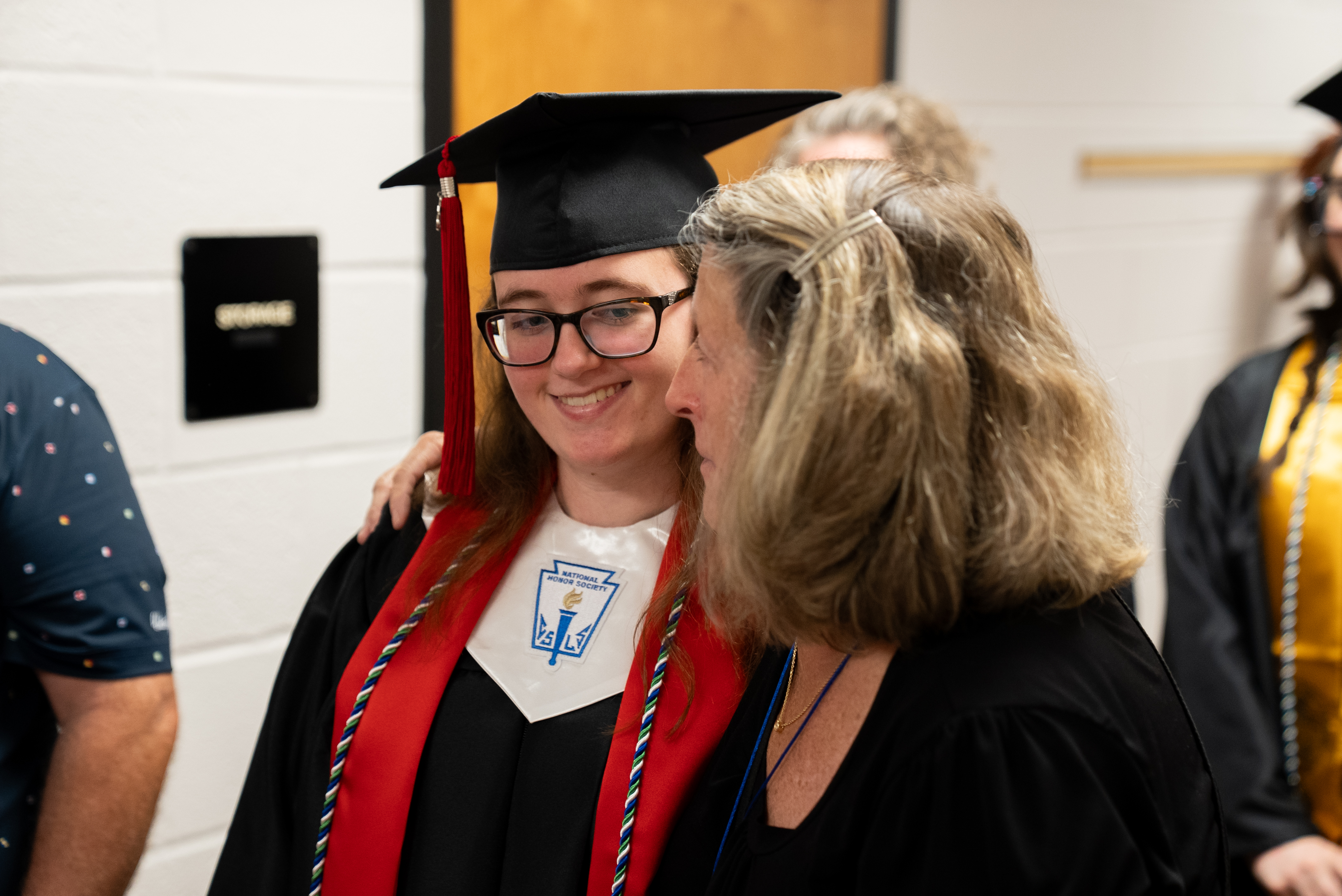 Woman with glasses and cap speaks with smiling woman in graduation robes. White walls and door in background.