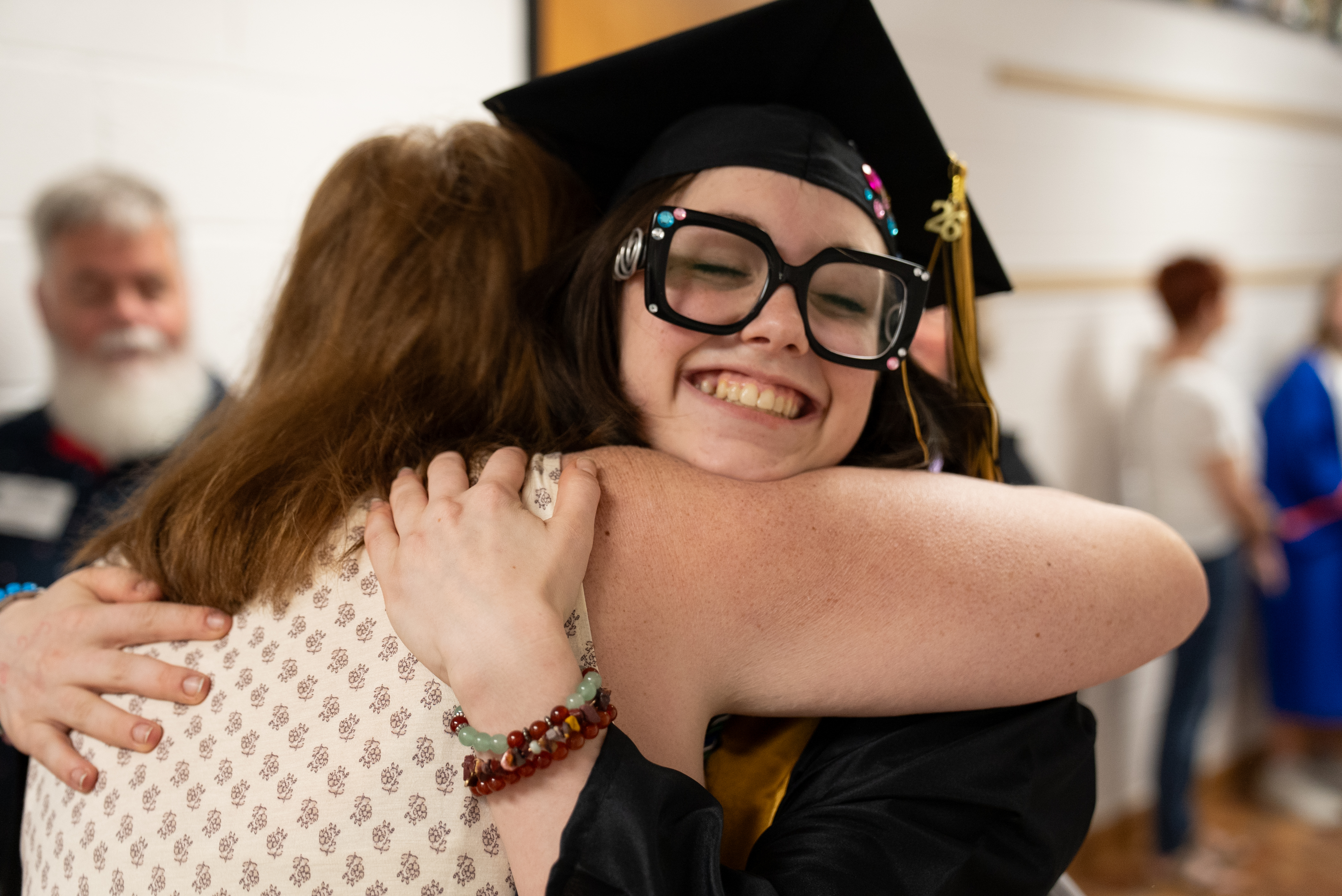 Woman in graduation gown and glasses hugging another person. Two other people stand in the background.