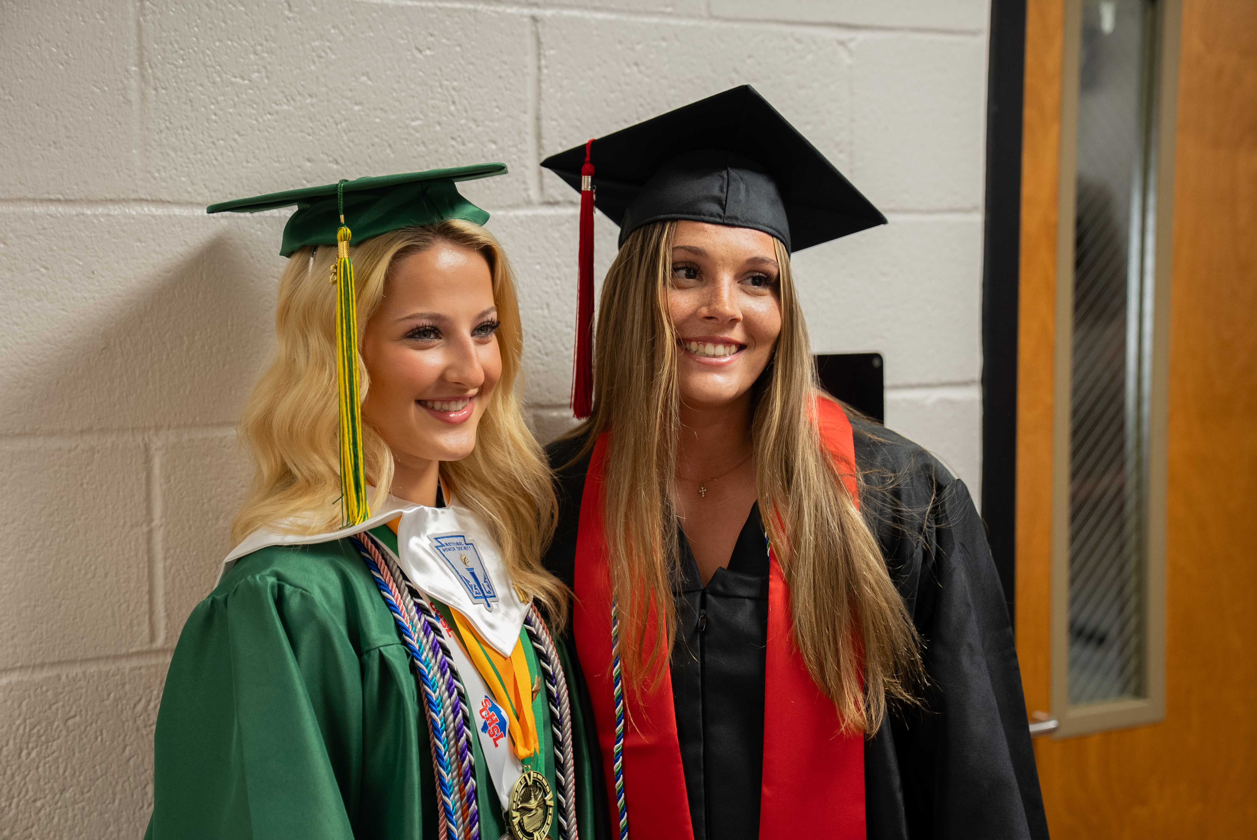 Two women in graduation gowns stand together, smiling. One wears green with medals, the other black with a red stole. Behind them, a white wall and an open door.
