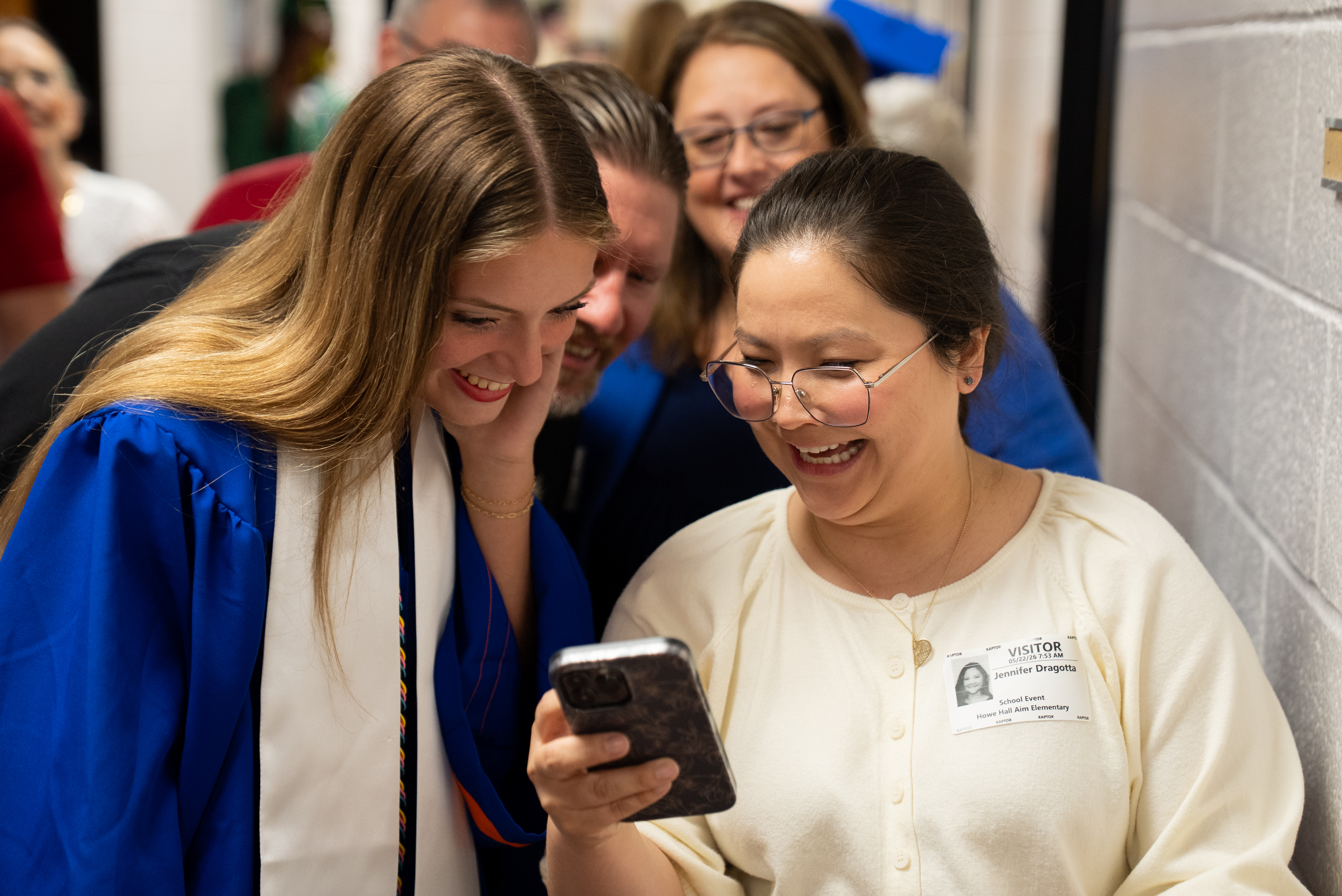 Two women, one in a graduation robe, smile and look at a cellphone. Behind them, people are blurred in a hallway.
