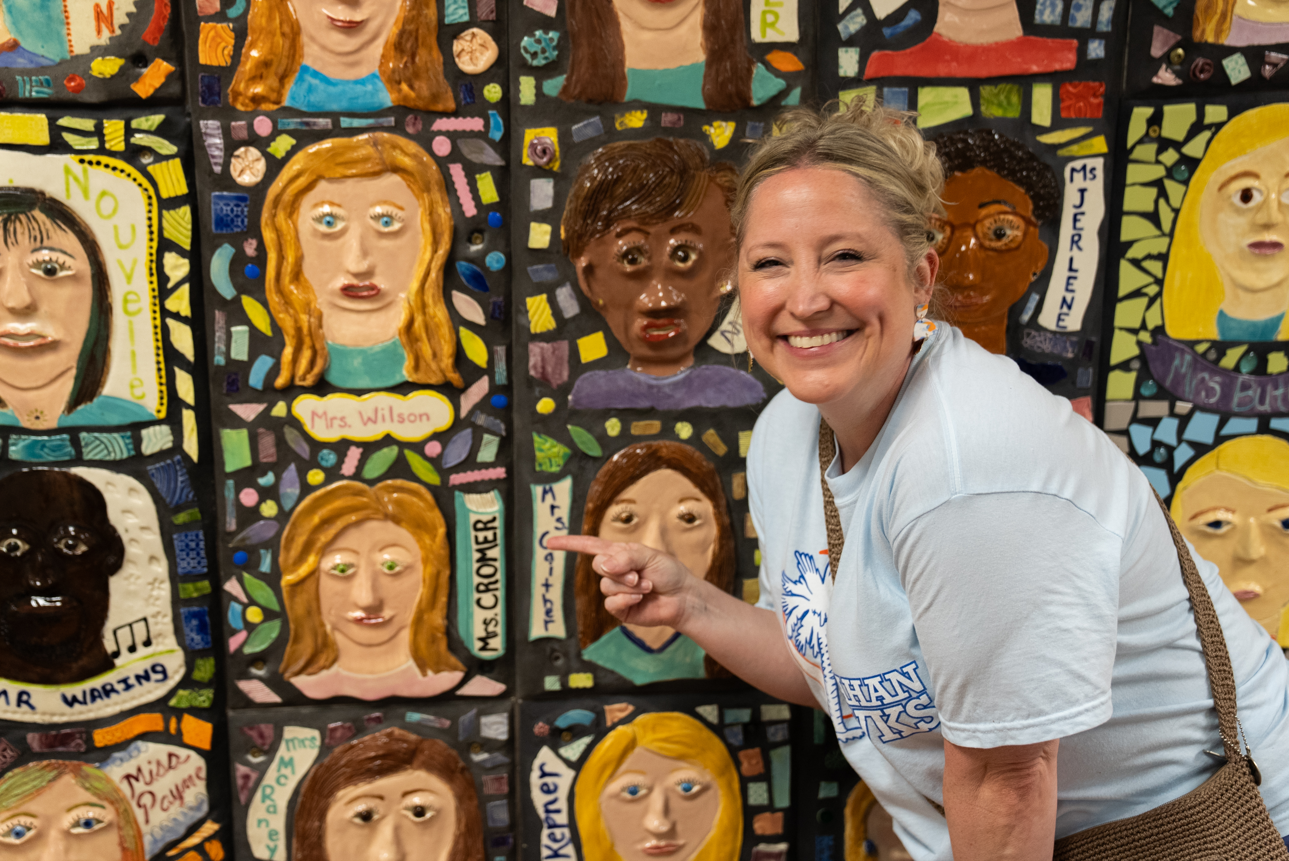 Woman with a bag smiles and points at a mosaic wall with different faces and names.