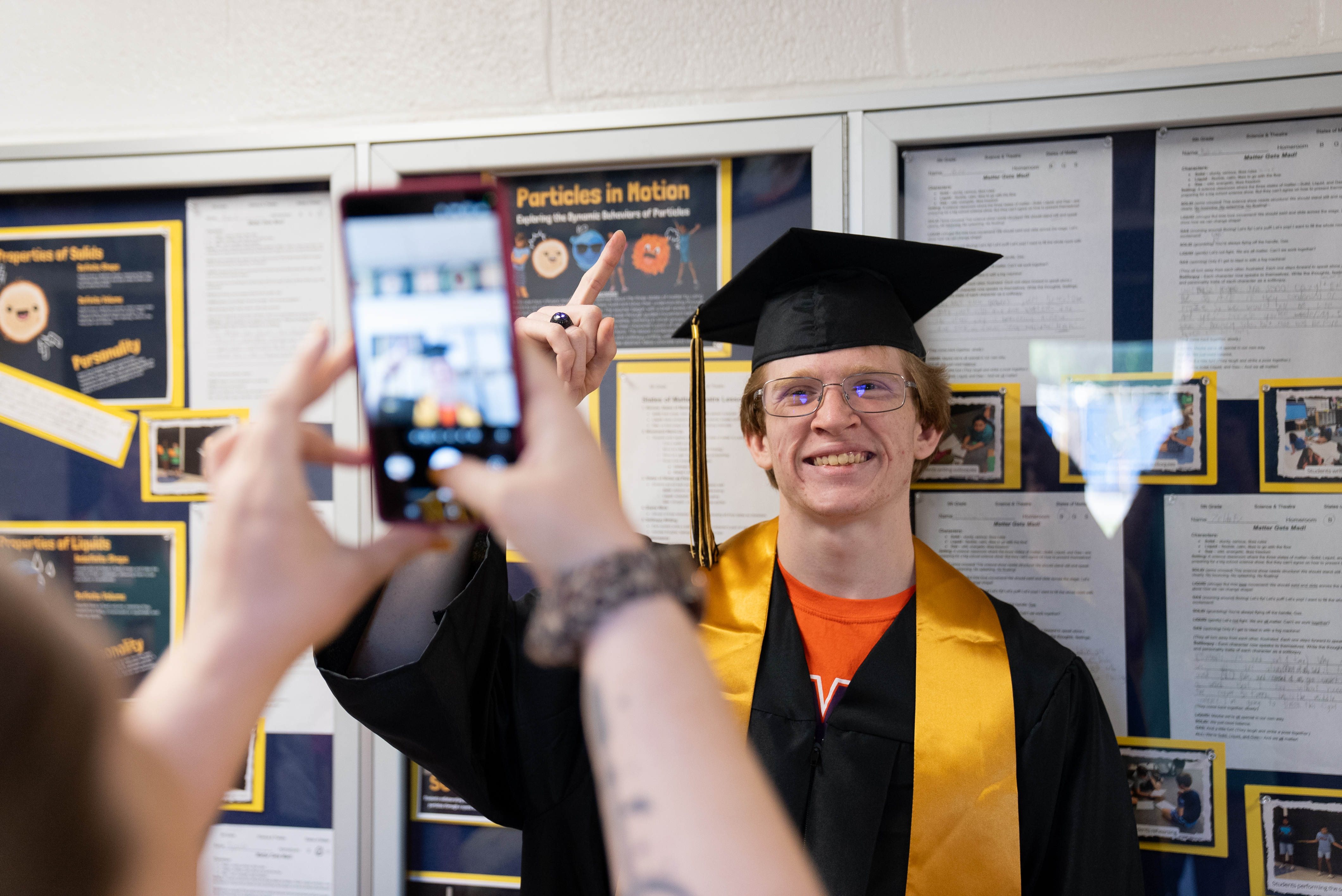 A person in a cap and gown takes a photo with a smartphone. Behind them, a wall displays various papers and pictures.