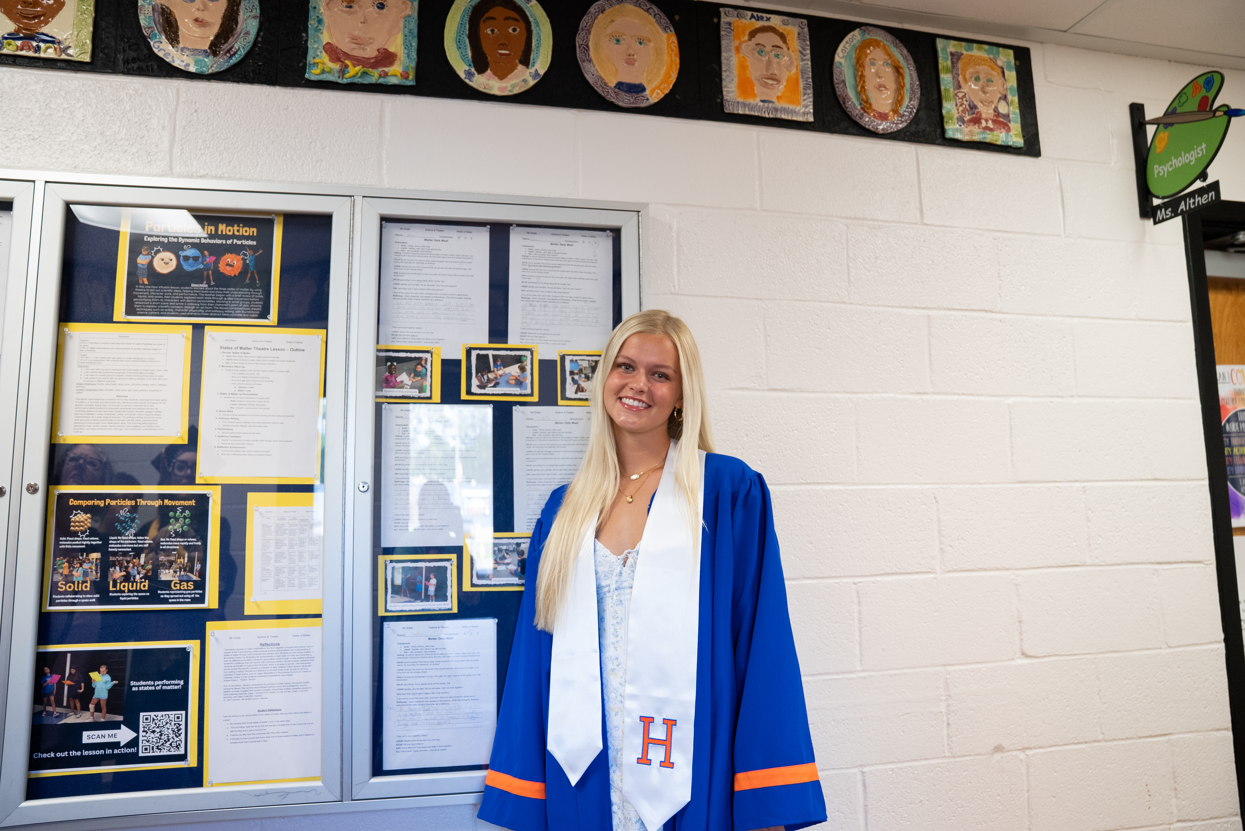 A woman with blonde hair stands in front of a bulletin board, wearing a blue and white graduation gown.