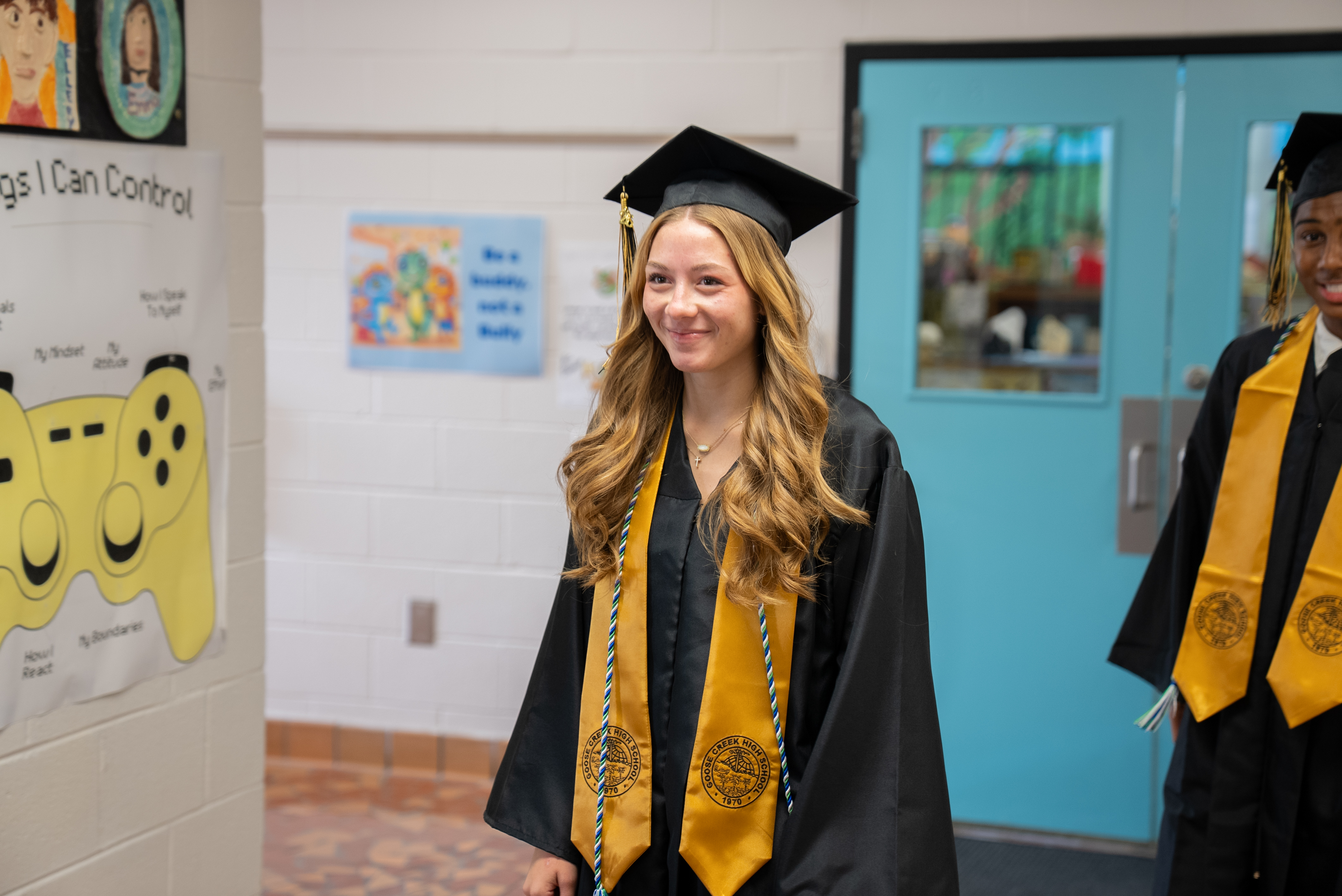 A woman in a graduation gown and cap smiles. A door with a window is behind her.