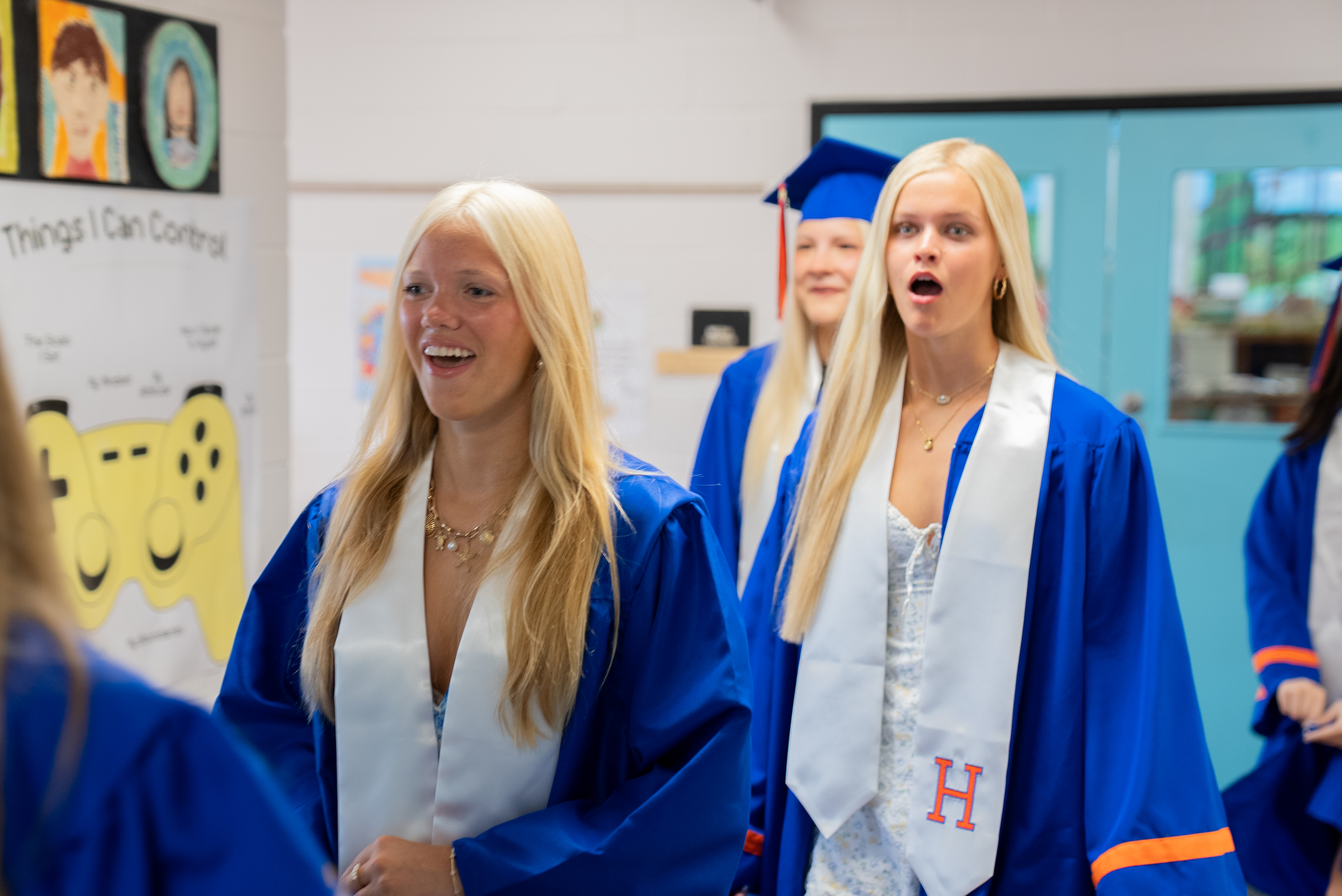 Graduates in blue gowns and white stoles, one with a sash reading "H", walk together, smiling.