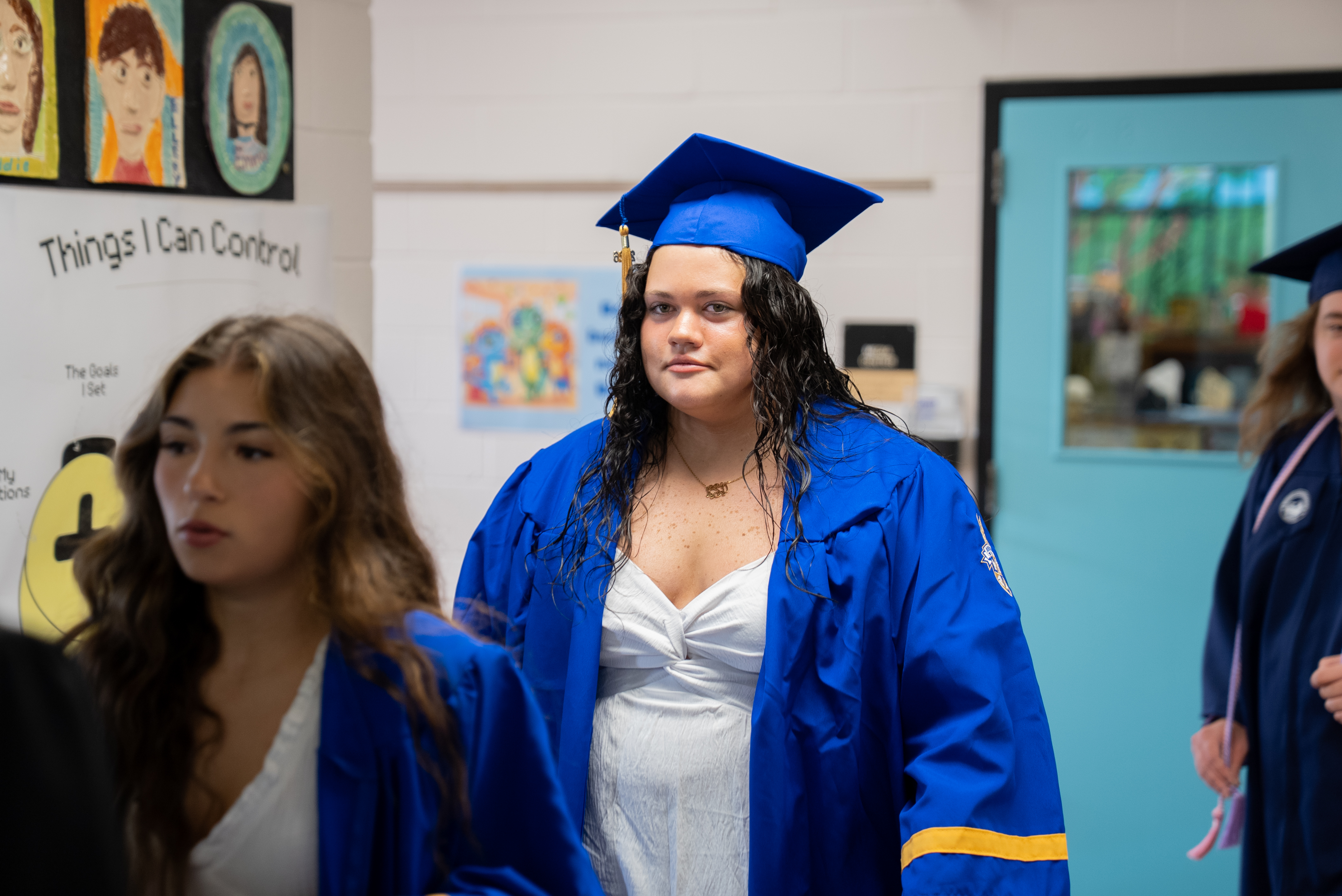 A woman in a blue graduation gown walks through a hallway with two other women in similar attire.