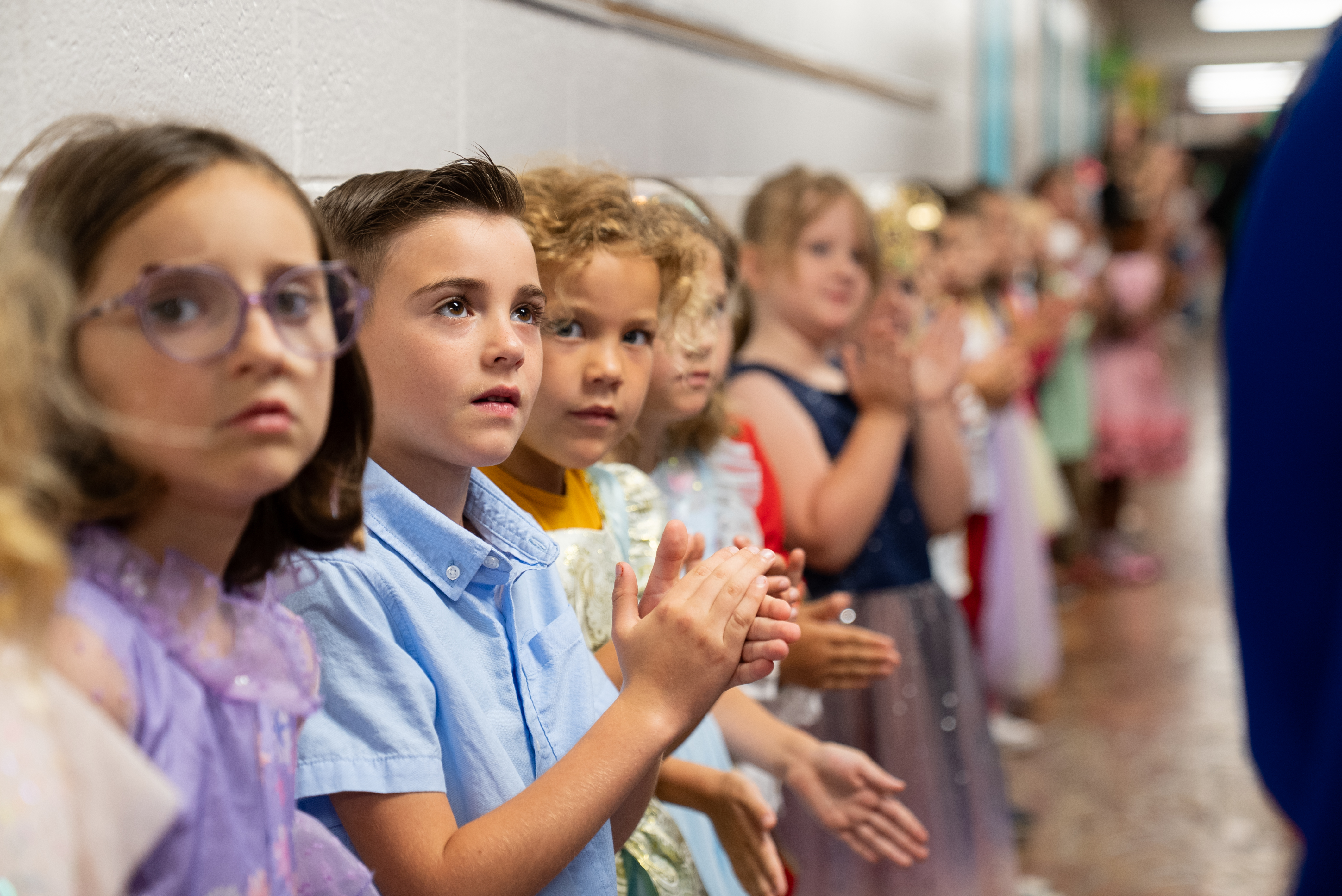 Group of children in a line, clapping. One child wears glasses. Background shows a hallway.