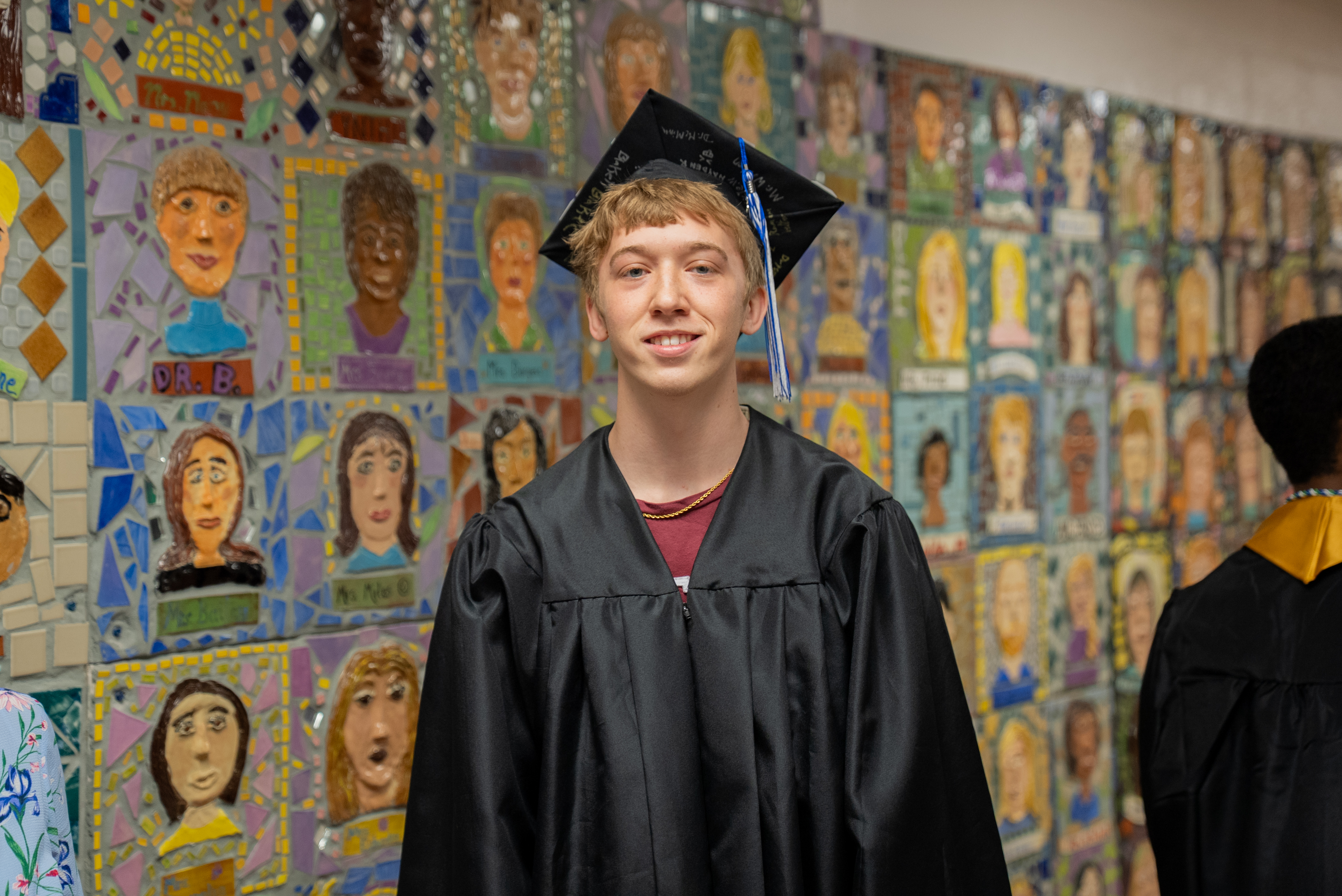 A young man in a black graduation gown stands in front of a colorful mosaic wall with many faces.