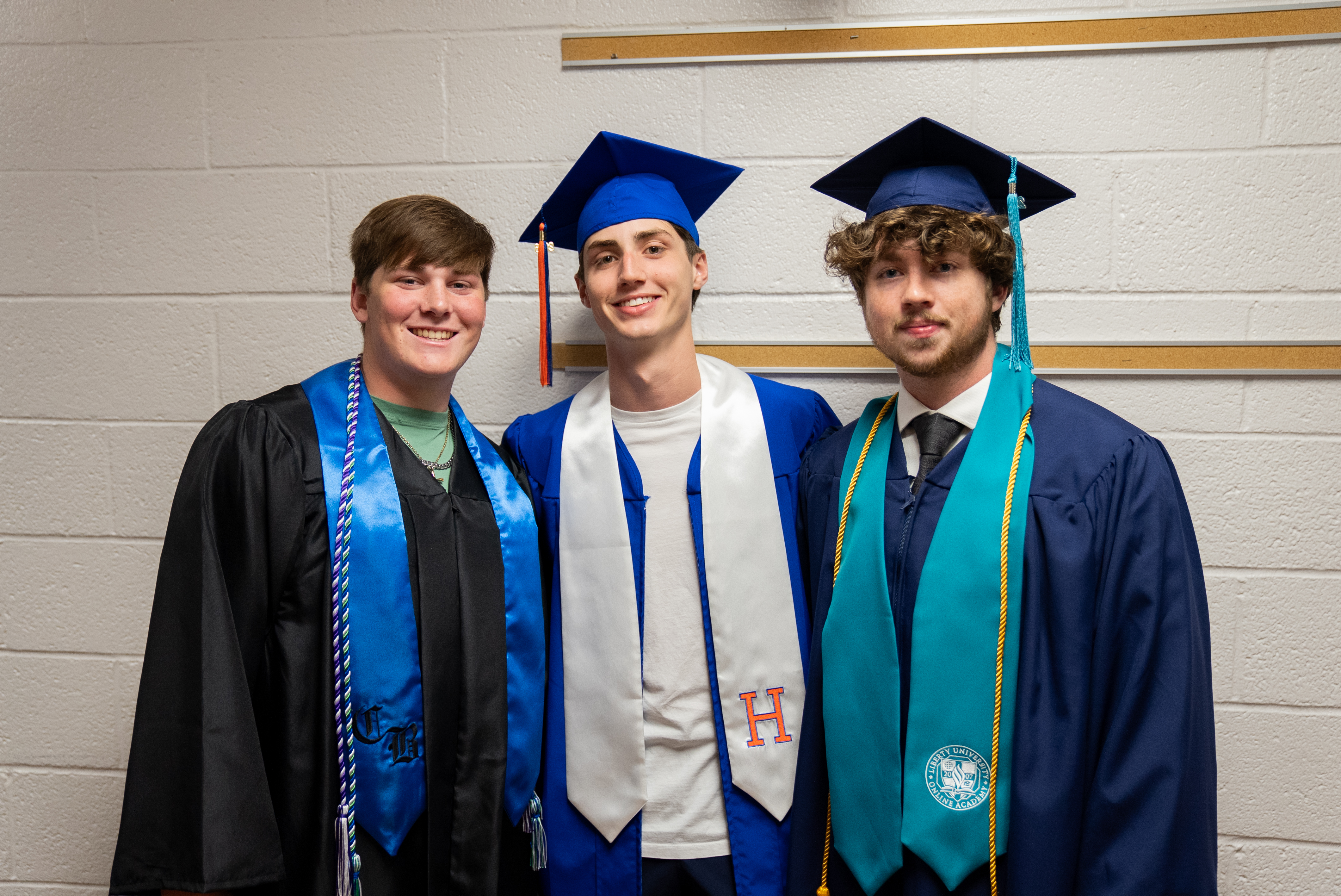 Three men in graduation gowns and caps pose for a photo in front of a white wall.