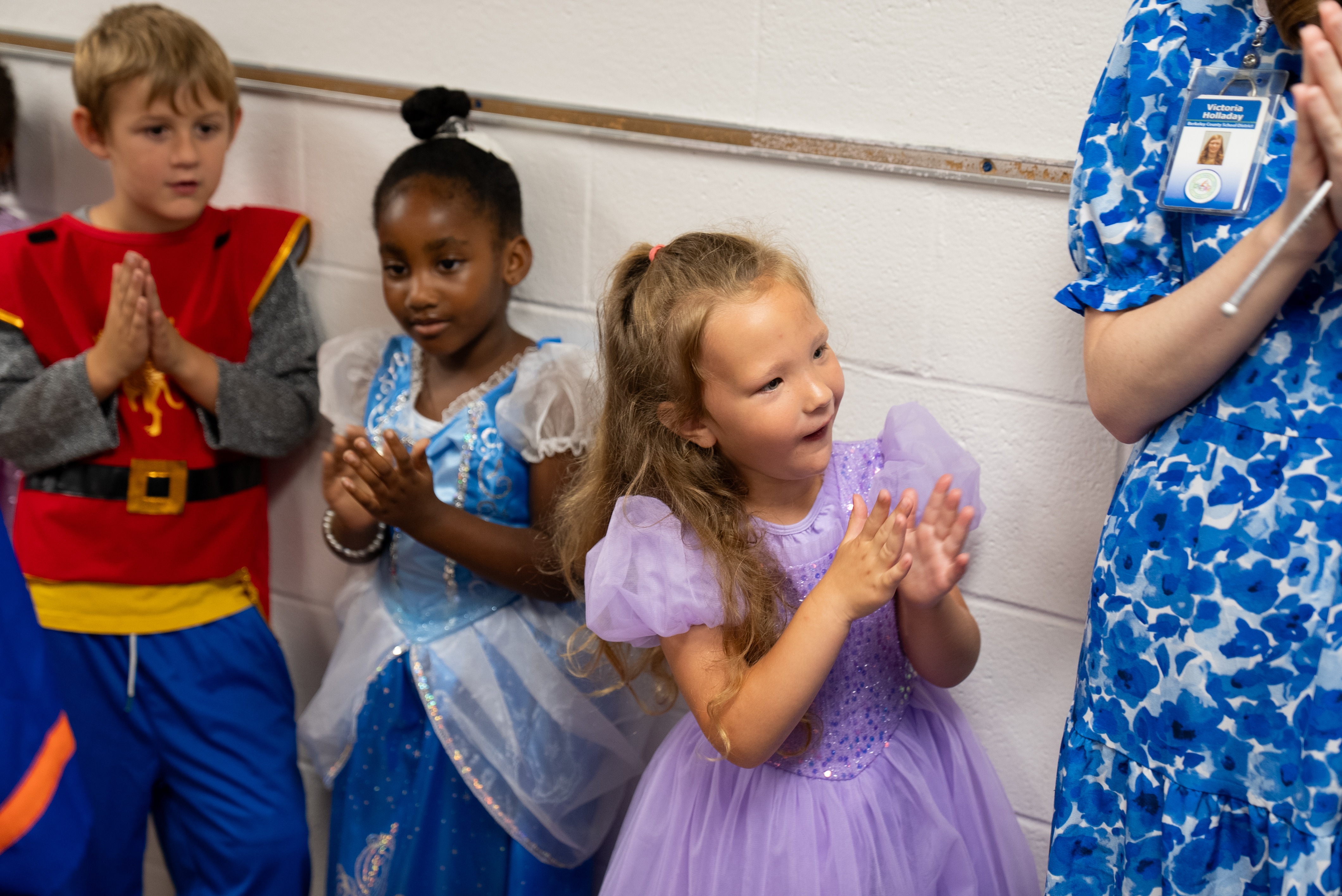 Several children in costumes, one girl in purple, a girl in a blue dress, a boy in red and blue, and a woman in blue.