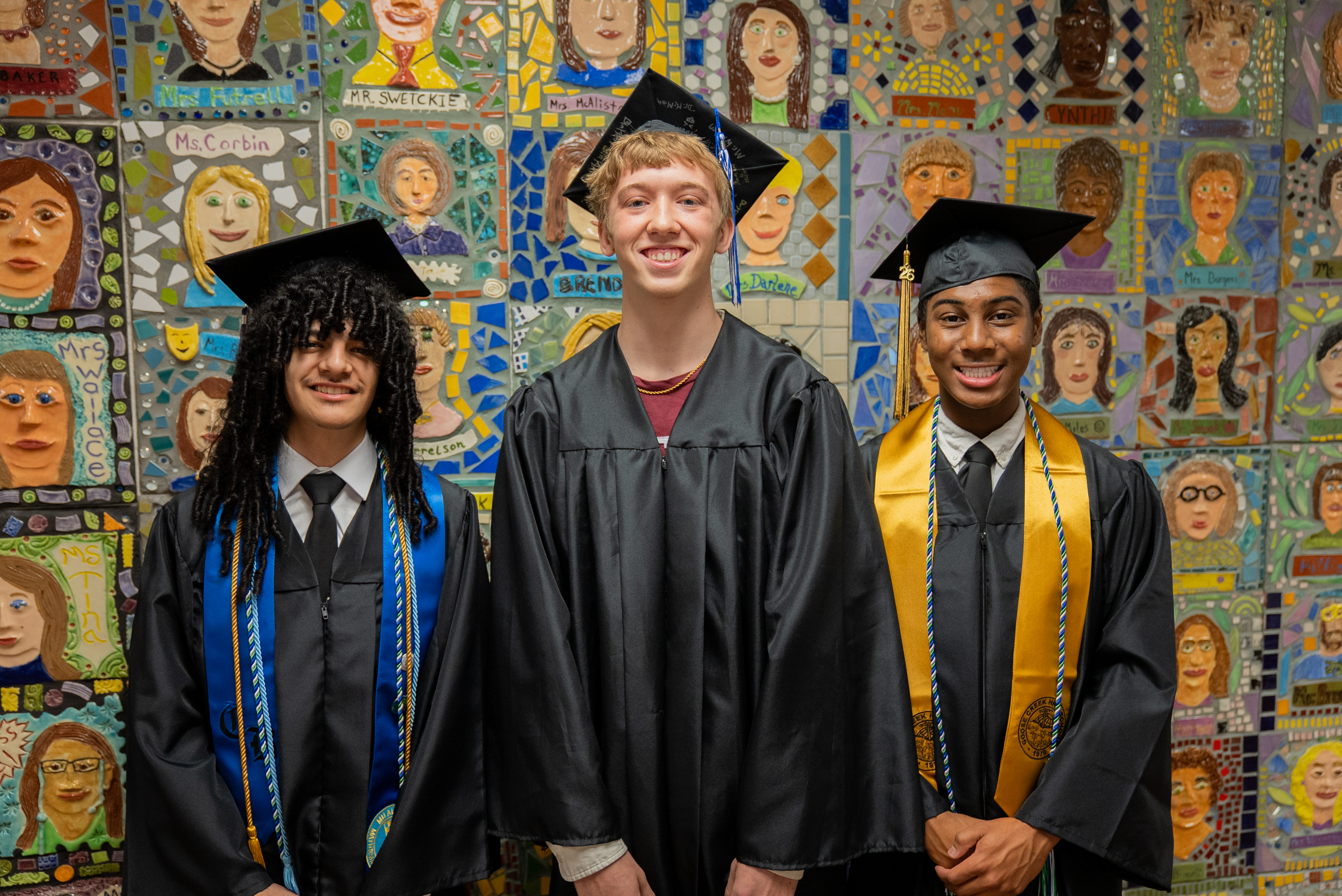 Three young men in graduation gowns and caps standing together in front of a wall decorated with mosaics.