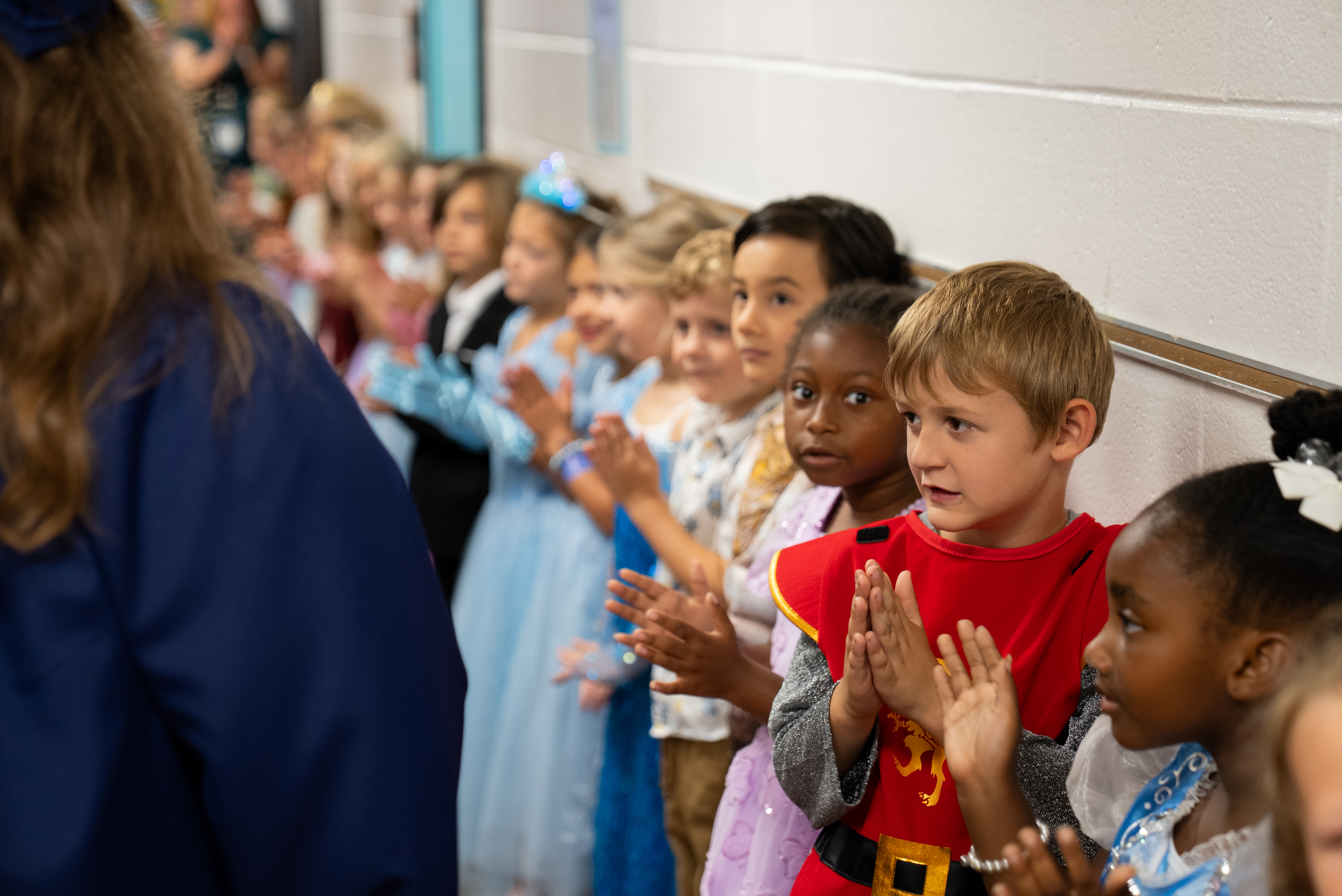 Kids in costumes, clapping in a line, with an adult at the front and a white wall in the background.