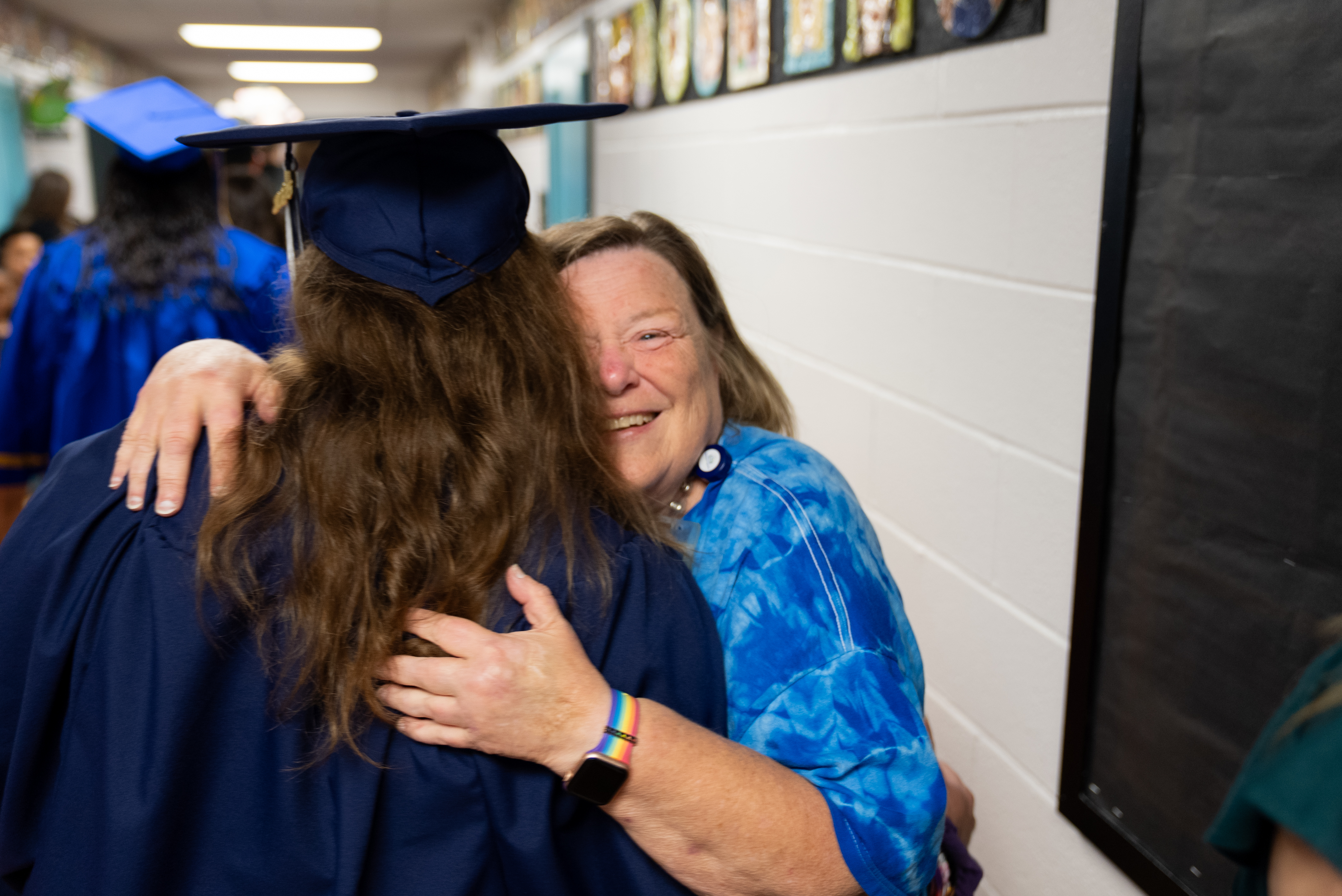 Woman wearing a graduation gown hugs a woman in a blue shirt, both smiling. Behind them, a hallway with a blackboard and picture frames on the wall.