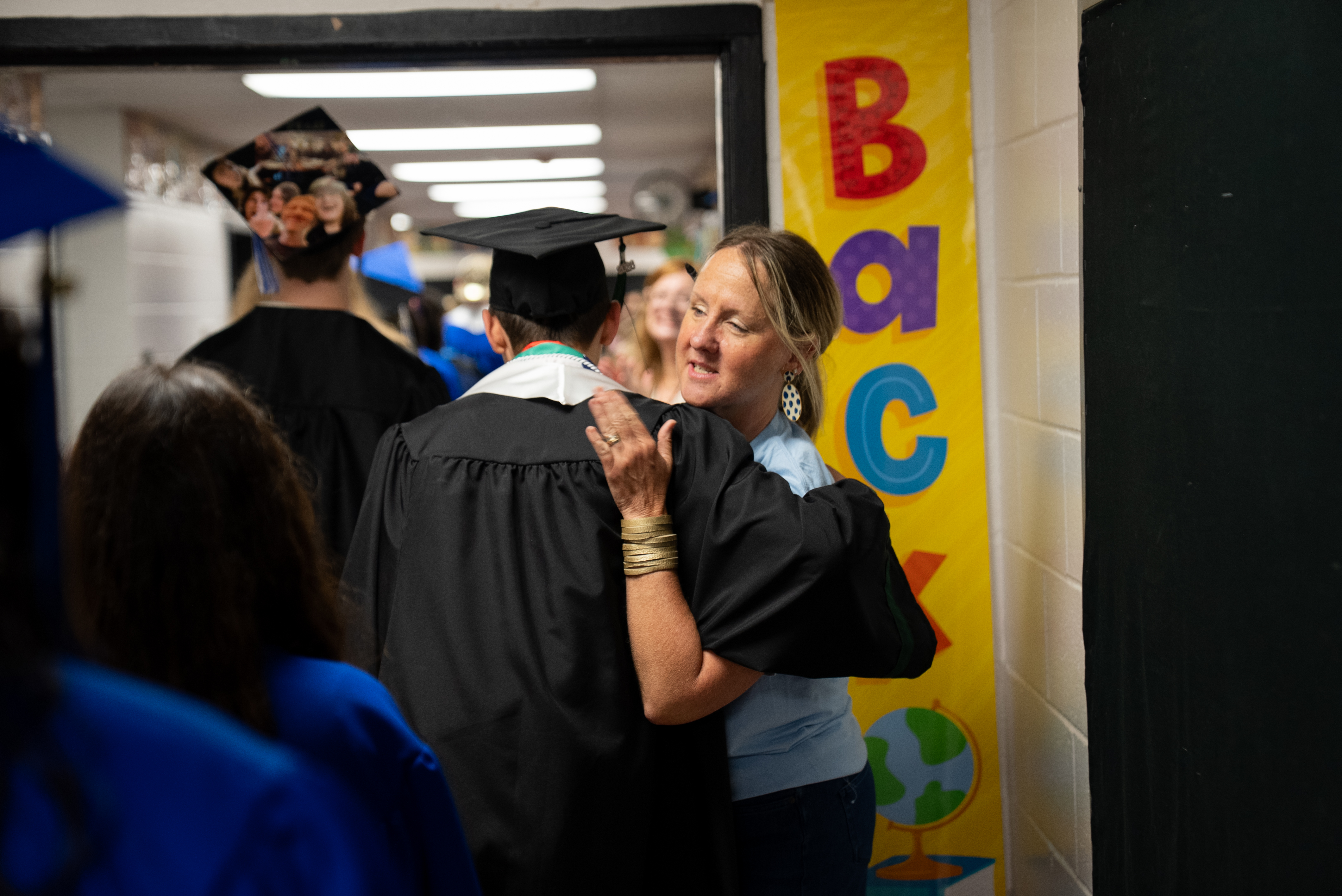 A woman hugs a graduate wearing a cap and gown in a corridor. The corridor has a yellow banner reading 'Back'.
