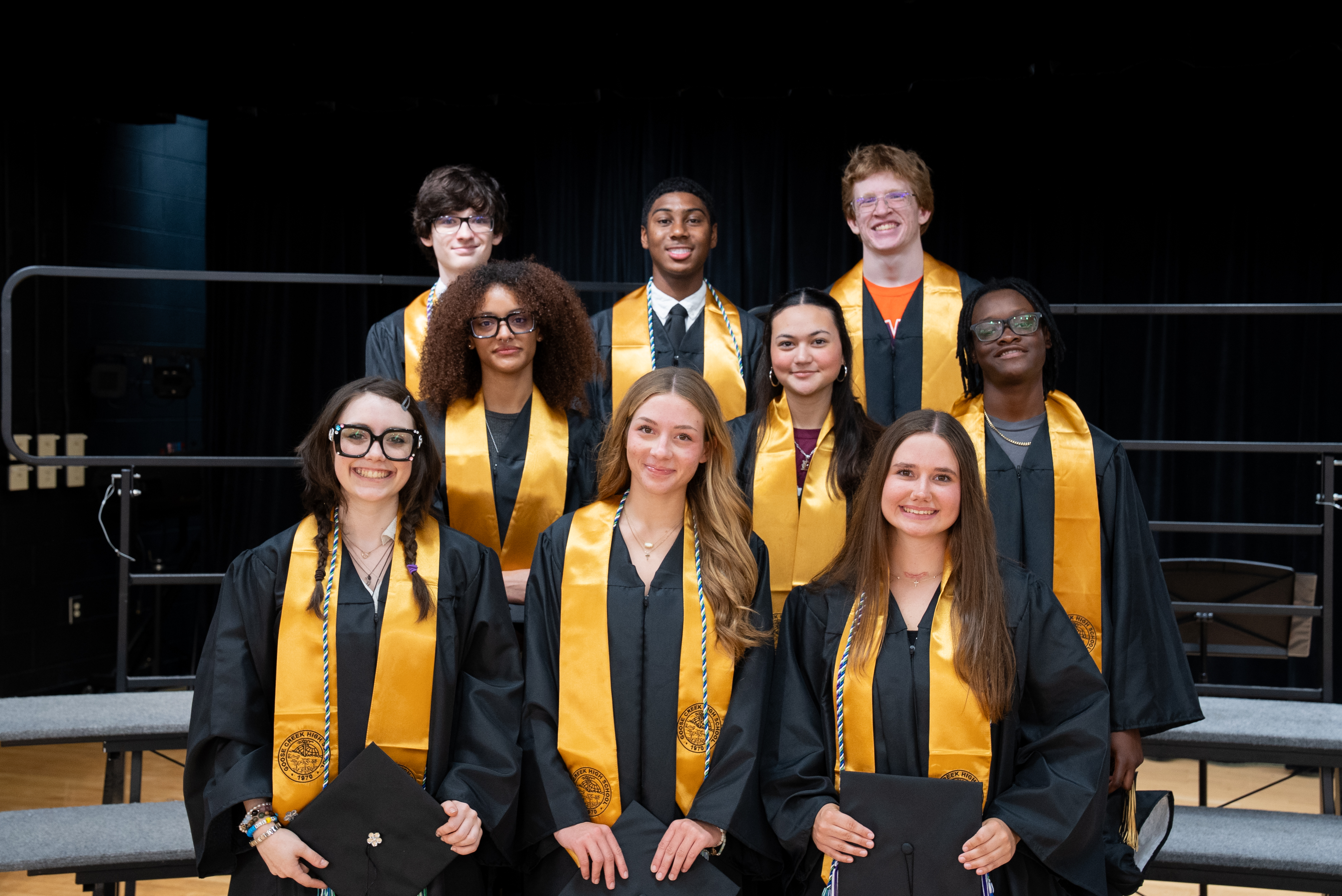 A group of students in graduation gowns and caps stands on a stage, smiling for a photo.