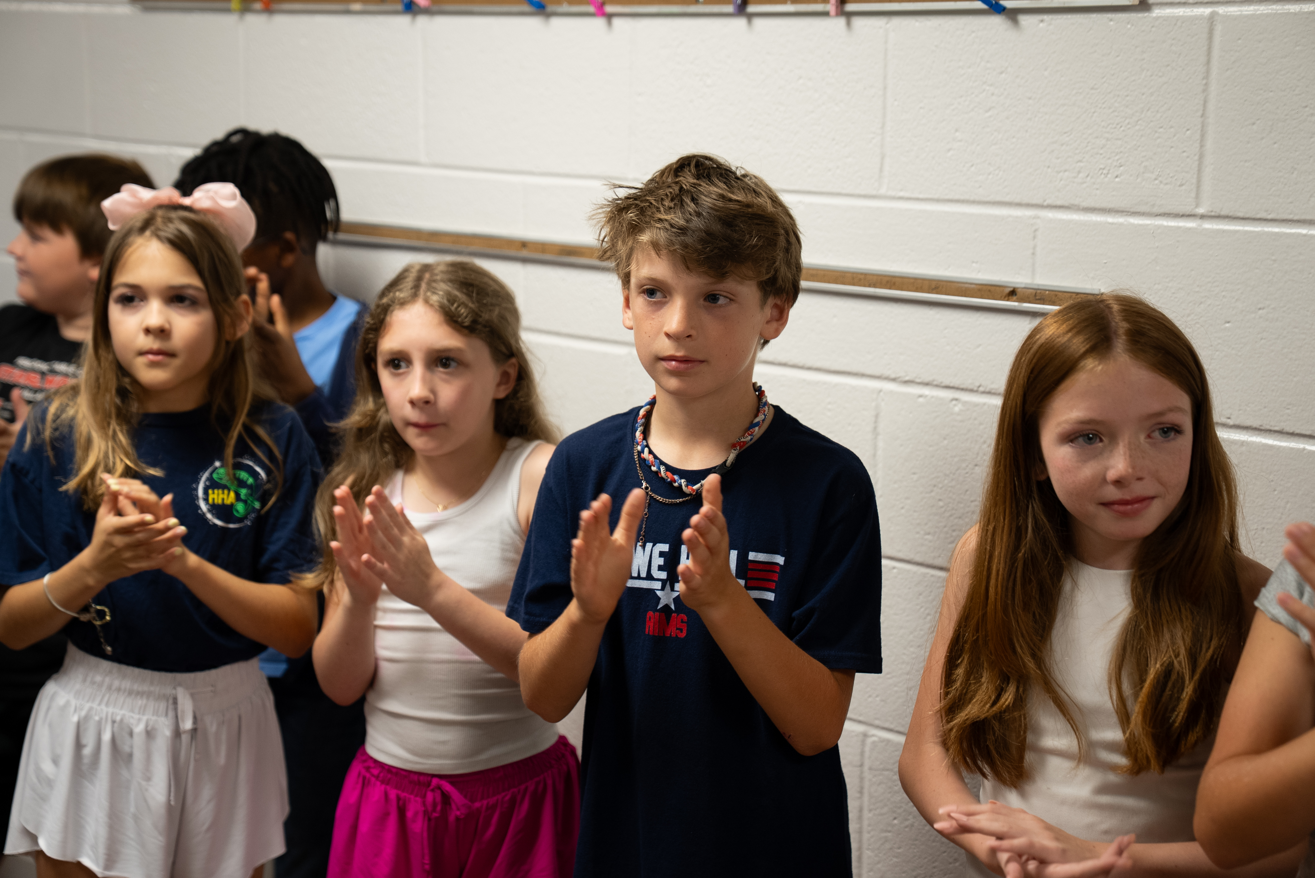 A group of children in a classroom. They are clapping and standing against a white wall.