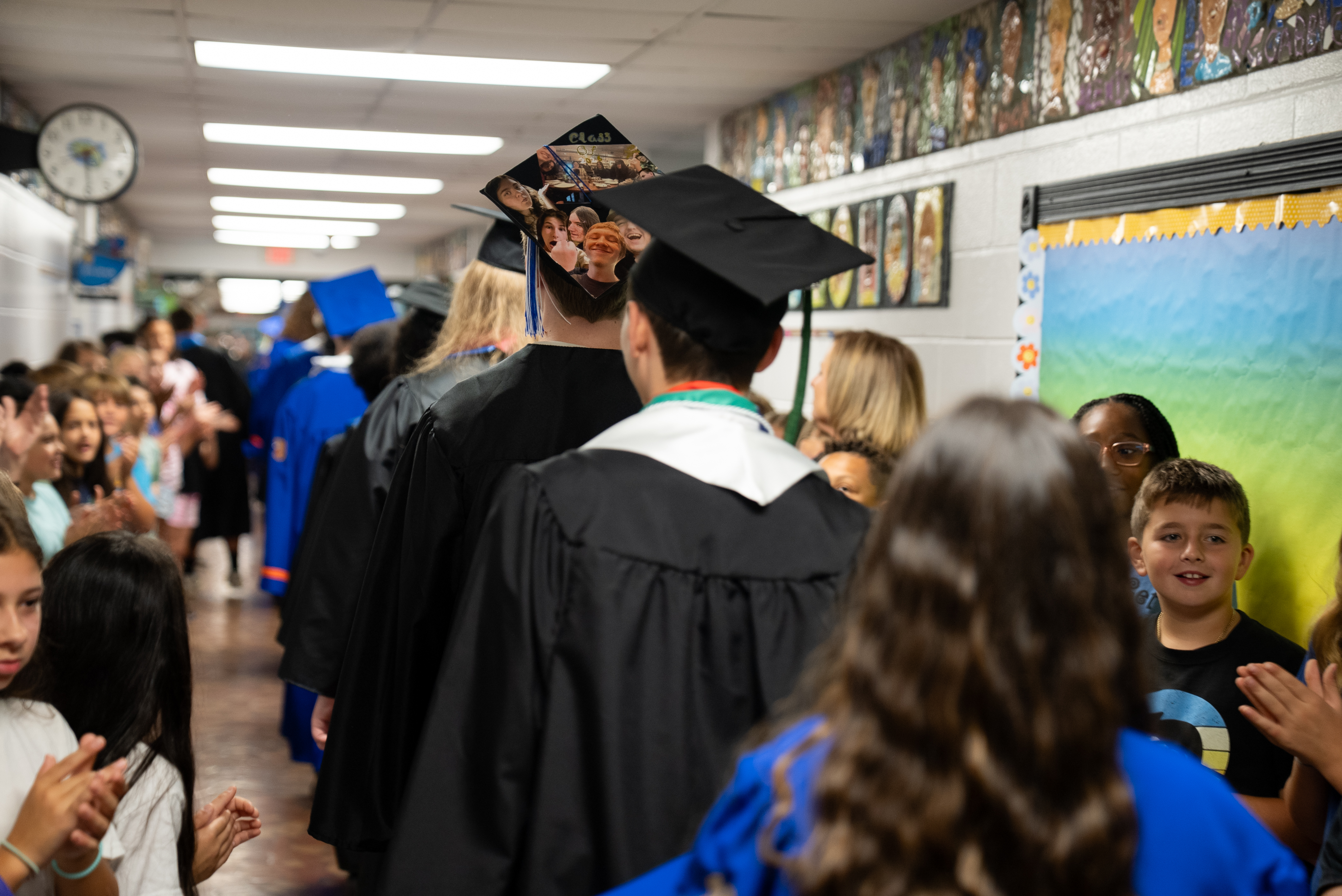 A corridor with people in caps and gowns. Some are walking, others are clapping. The walls have artwork and a clock.
