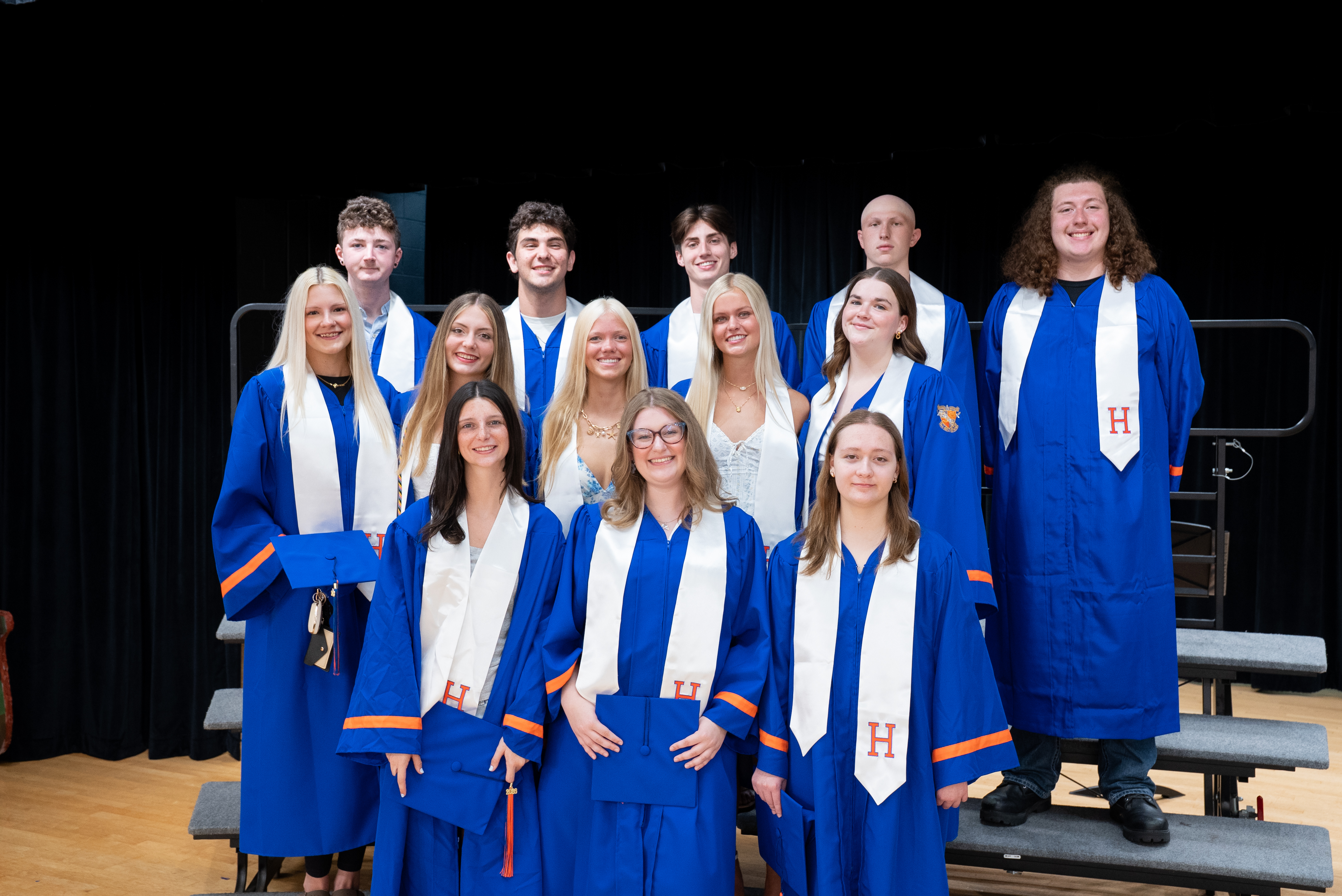 A group of students wearing blue and white graduation gowns with the letter H, standing on a stage.